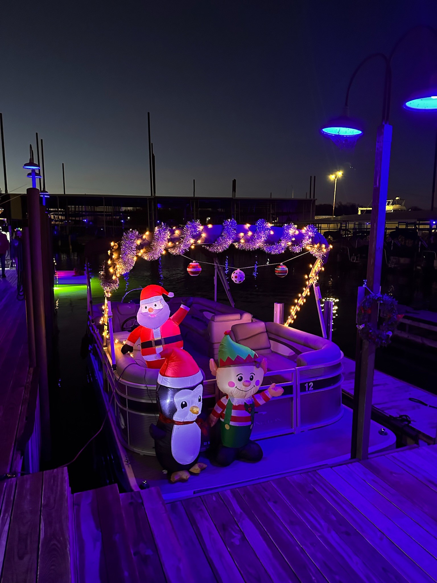 A festively decorated boat is docked at twilight, adorned with glowing fairy lights, inflatable Christmas figures, and ornaments, creating a cheerful holiday atmosphere.