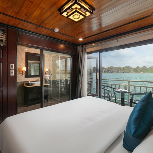 Luksuriøst værelse med en stor seng, glansfuldt træloft, skydedøre i glas, der åbner ud til en balkon med udsigt over havet og klipperne.