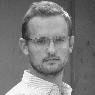 A person wearing glasses and a white shirt standing against the wall.