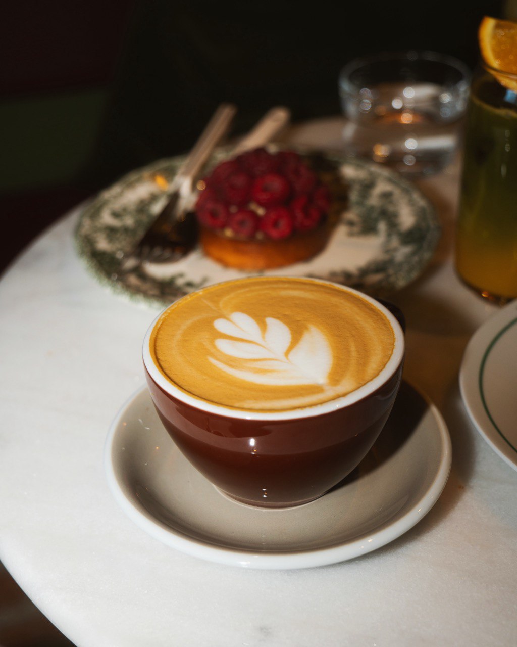 A beautifully crafted latte with intricate leaf art in a brown cup is placed on a saucer on a marble table, accompanied by a raspberry-topped dessert in the background, highlighting a cozy cafe ambiance.