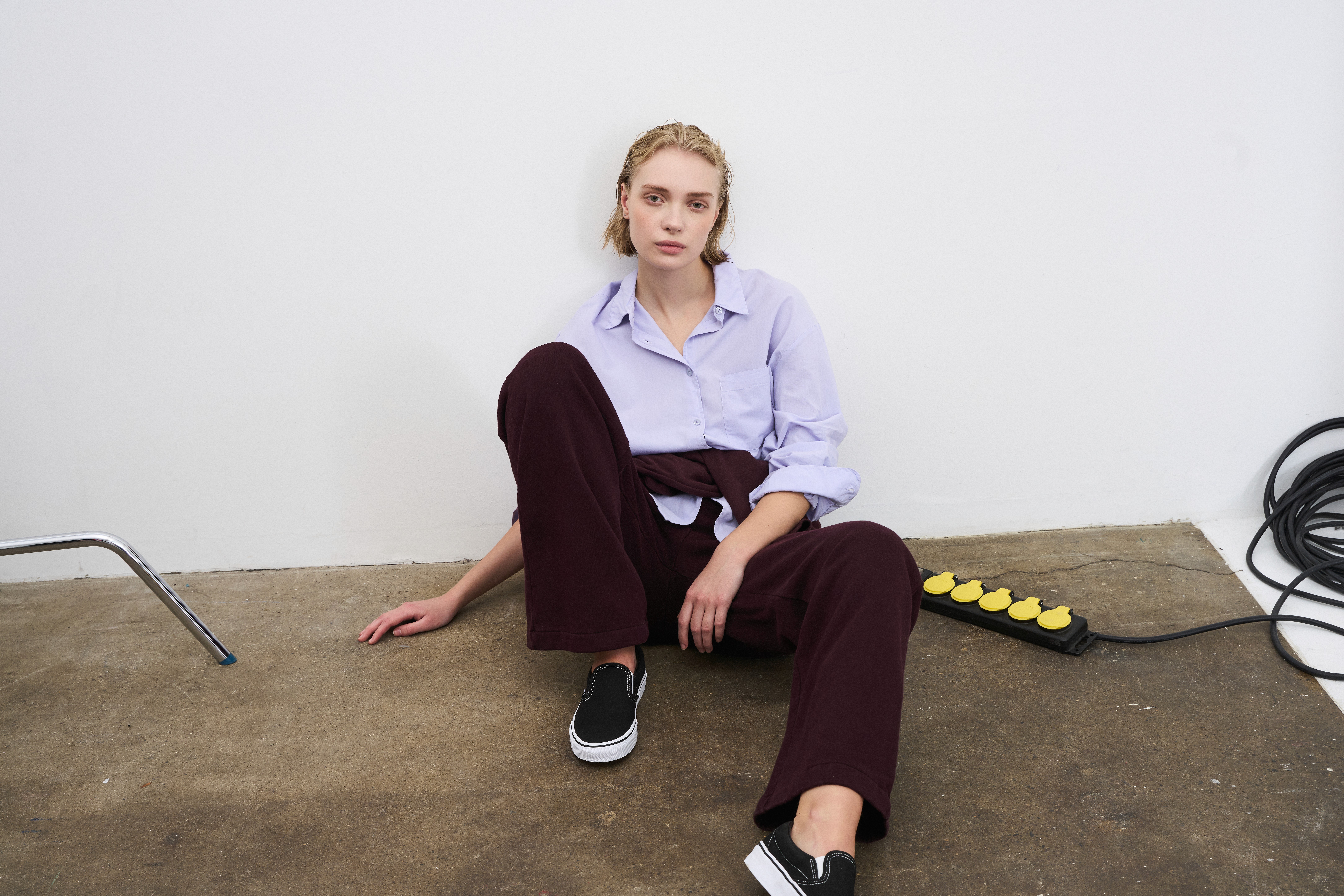 Dans le lookbook Colorful Standard, la mannequin porte un pantalon bordeaux et une chemise mauve, assise par terre