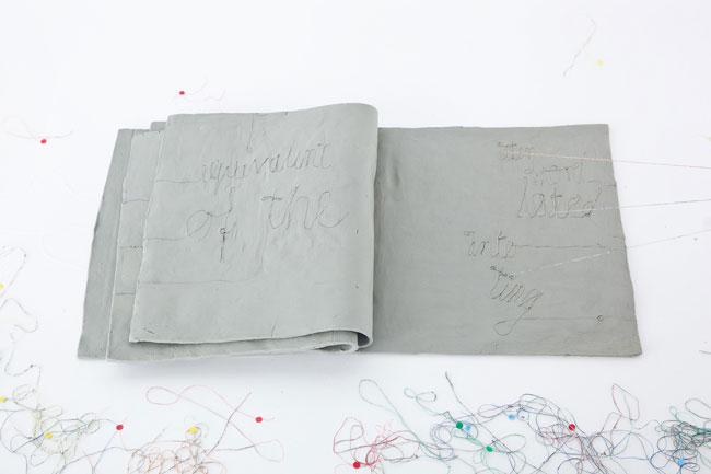 The image shows an open book with a gray cover, resting on a white surface. The pages have handwritten text in black ink, including "separative of the" on the left page and "wasn't liked" on the right page. Scattered around the book are colorful strings of yarn in various colors, including red, yellow, blue, and green. The strings extend beyond the edges of the book and lie on the surface around it. The white background provides a stark contrast to the gray book and the colorful yarn.