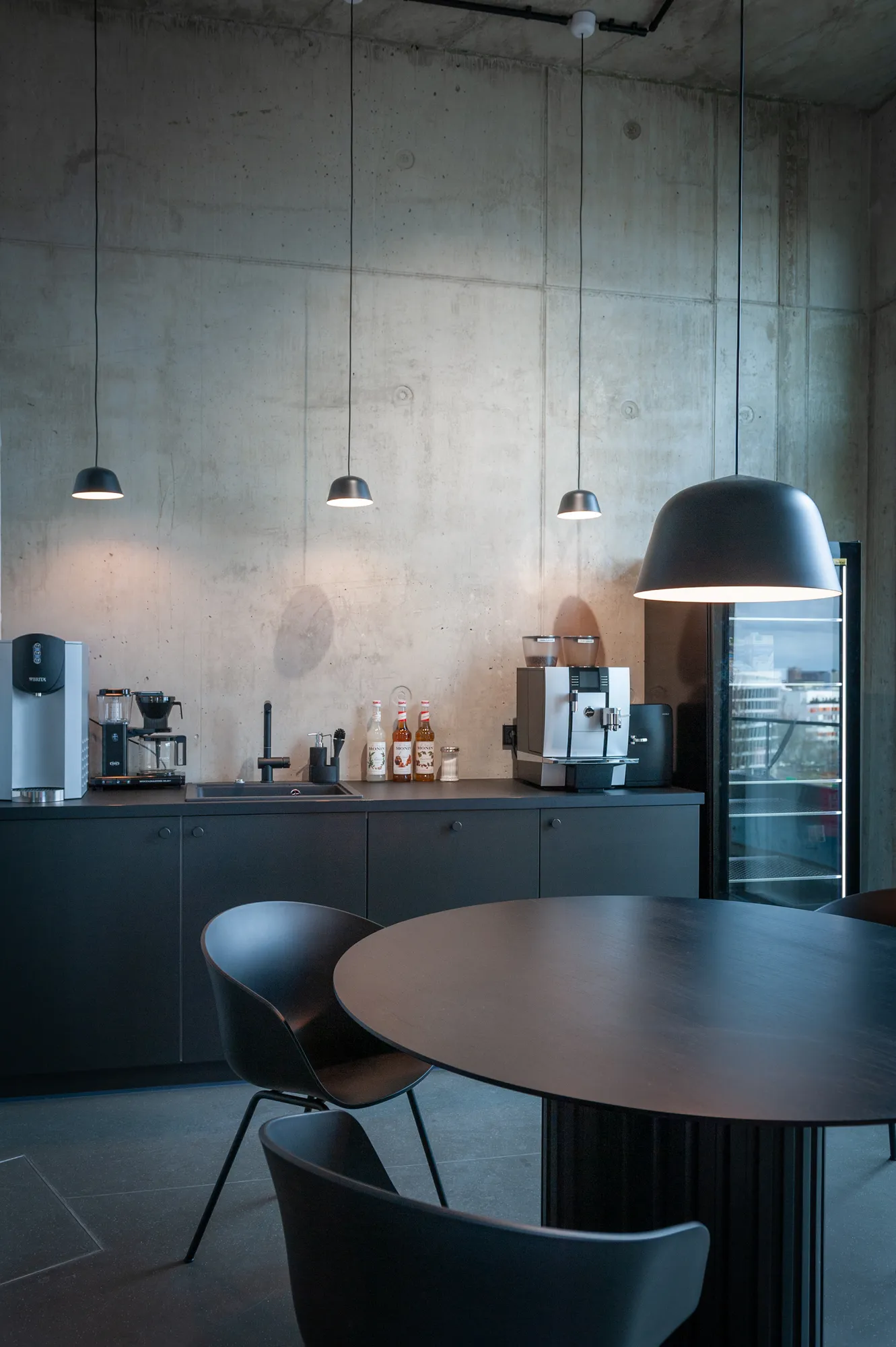Concrete-walled kitchen area with coffee machines, fridge and round dining table with chairs.