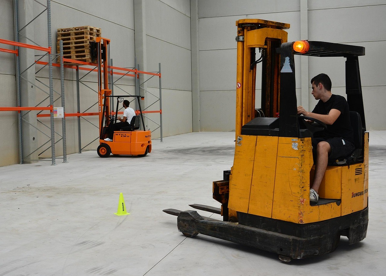 Two forklift operators in a warehouse, one on an orange forklift stacking pallets on shelving, the other on a yellow forklift.