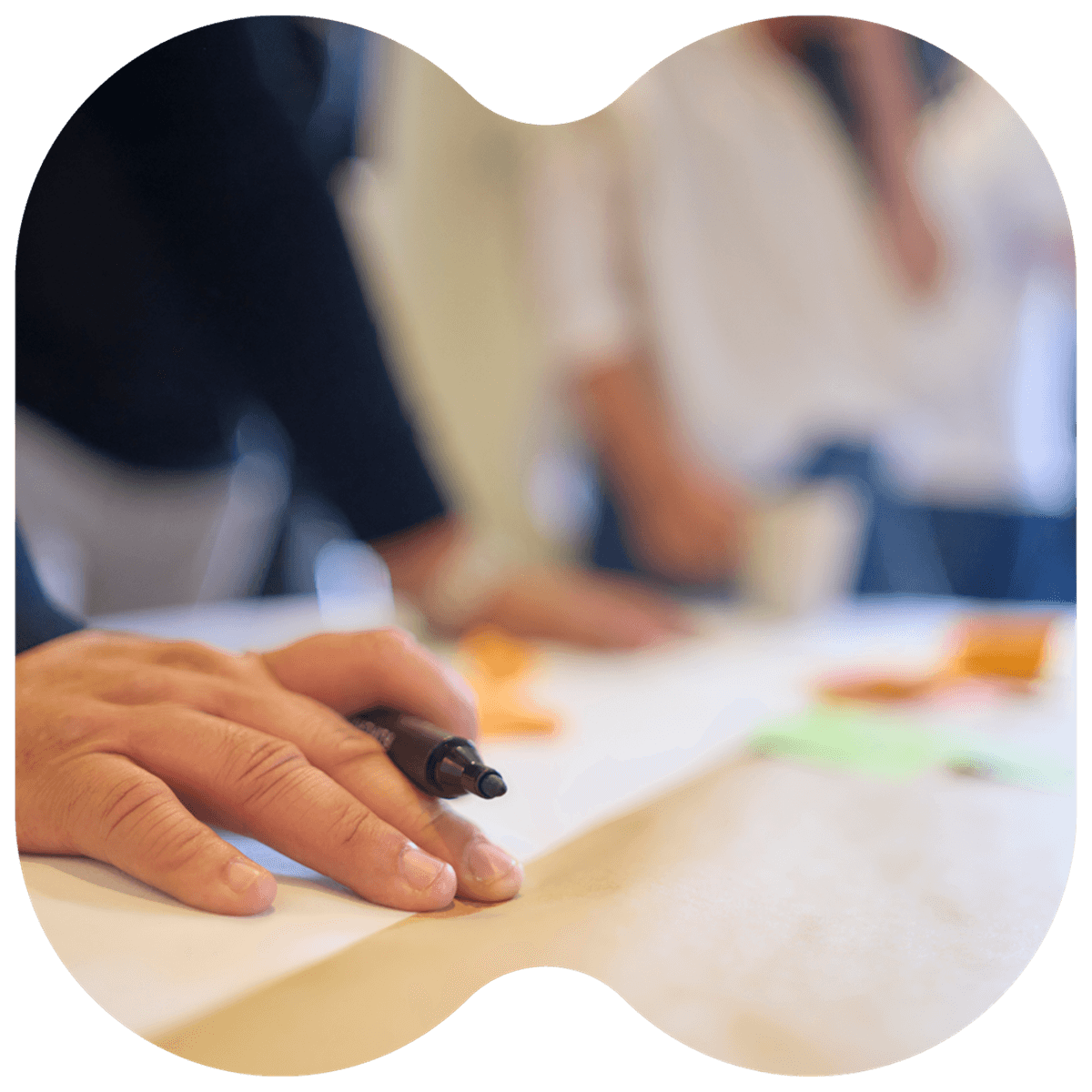 Close-up of a person's hand holding a pen with others in the background, likely during a meeting or brainstorming session.