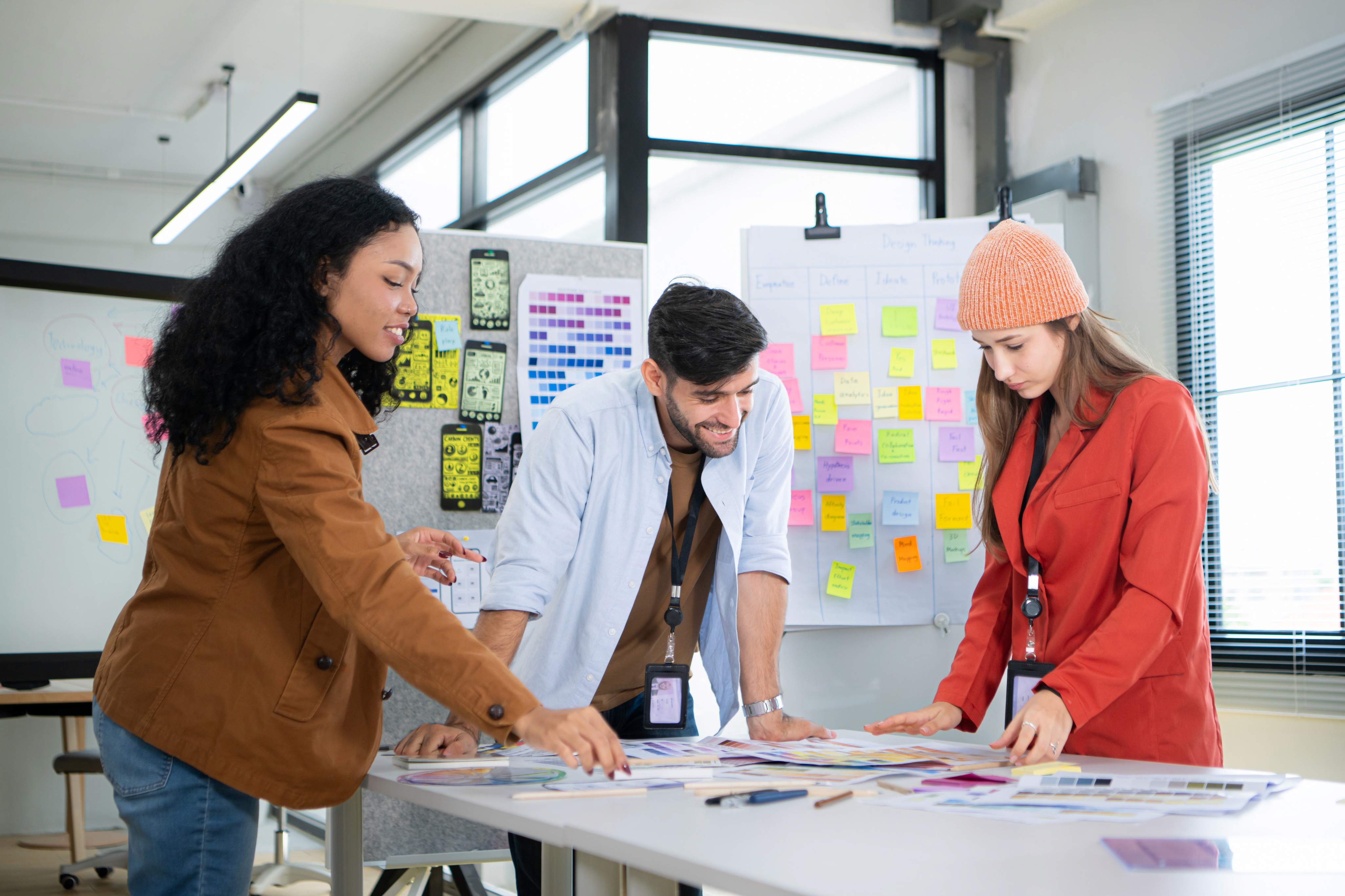 Three young designers collaborating at a table in front of colorful sticky note boards.