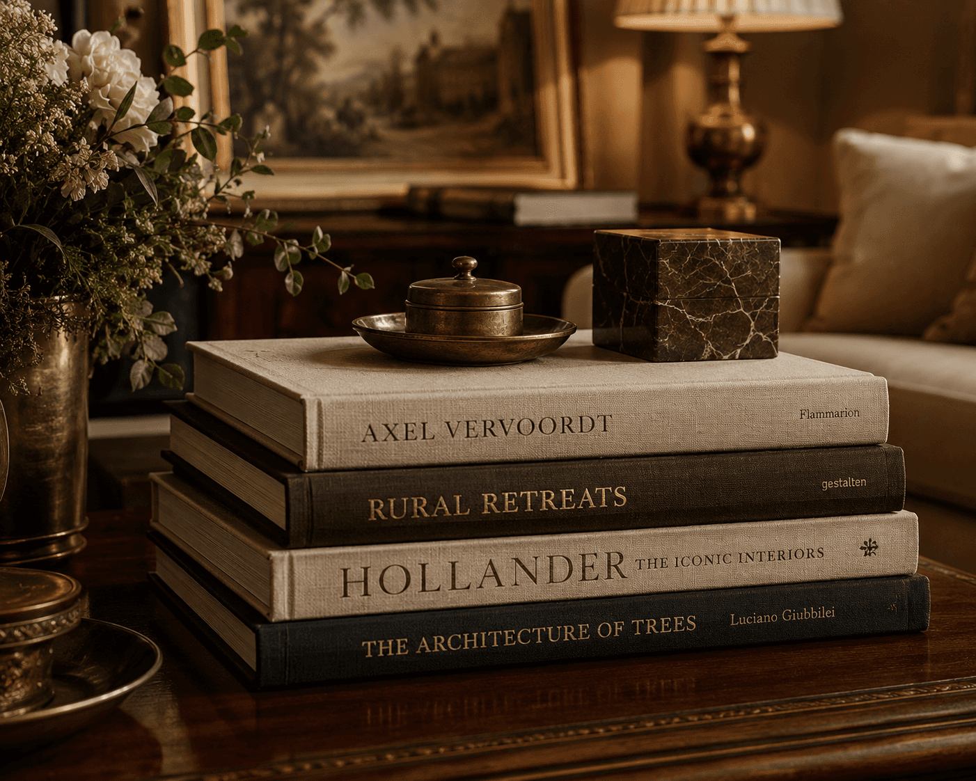 A stack of four hardcover design and architecture coffee table books styled on a polished dark wood surface. A brass dish with a lid and a brown marbled square box sit atop the books. In the background, a brass vase with white flowers, a warm lamp, and framed art add to the classic interior decor aesthetic.