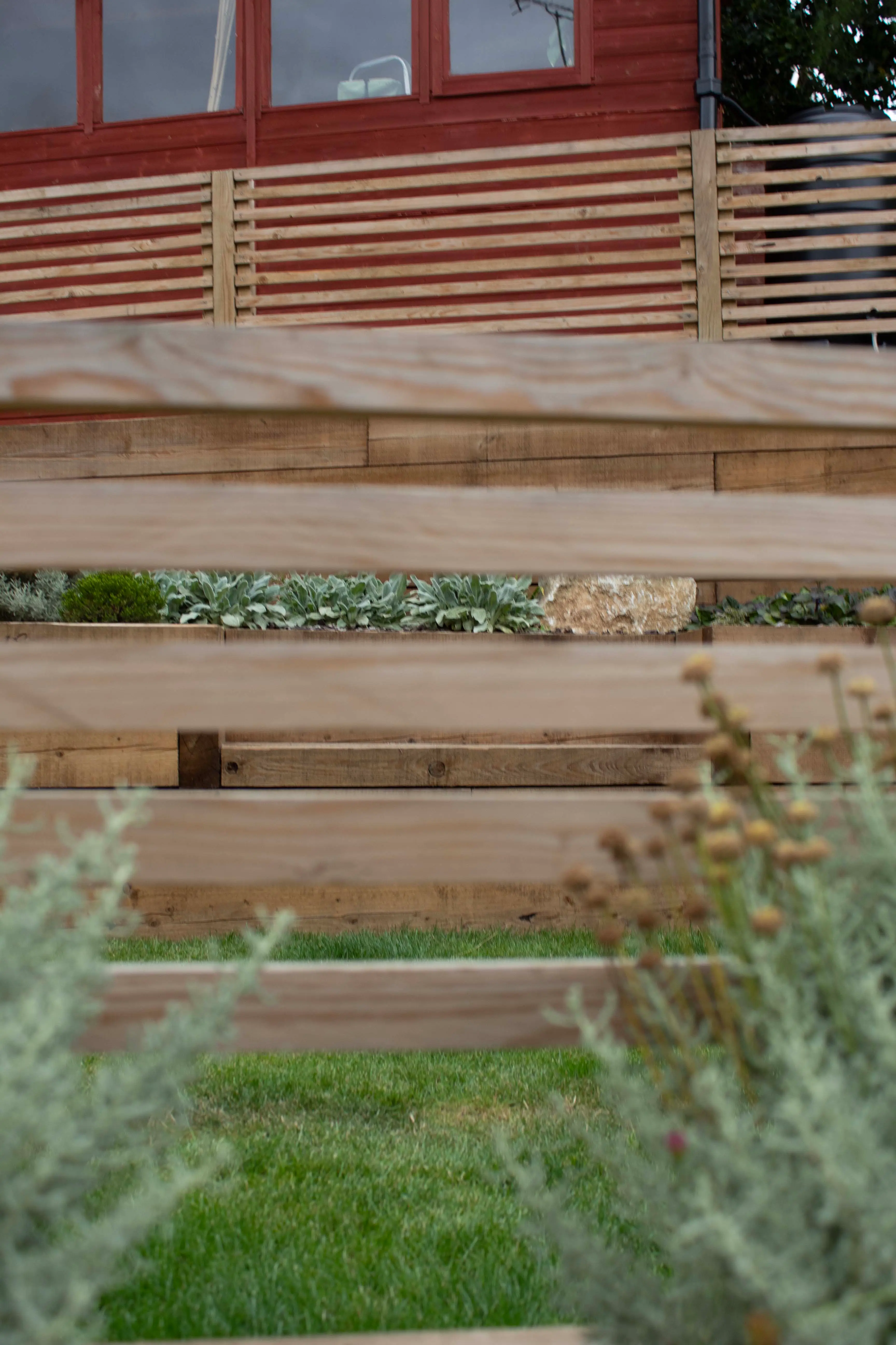 Close-up of wooden planks with green plants nearby, set against a background of a red building.