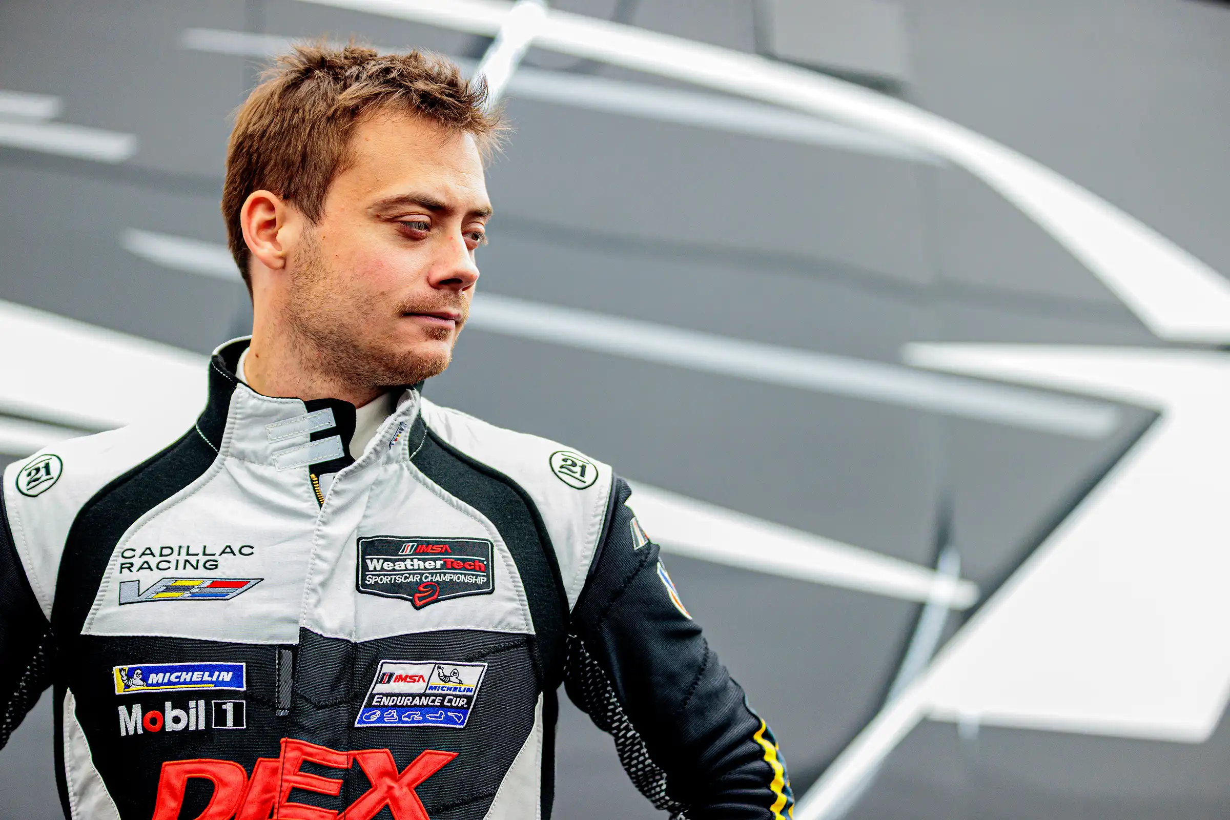 Cadillac Racing driver, Louis Delétraz, stands confidently in front of a gray patterned wall, looking to the right.