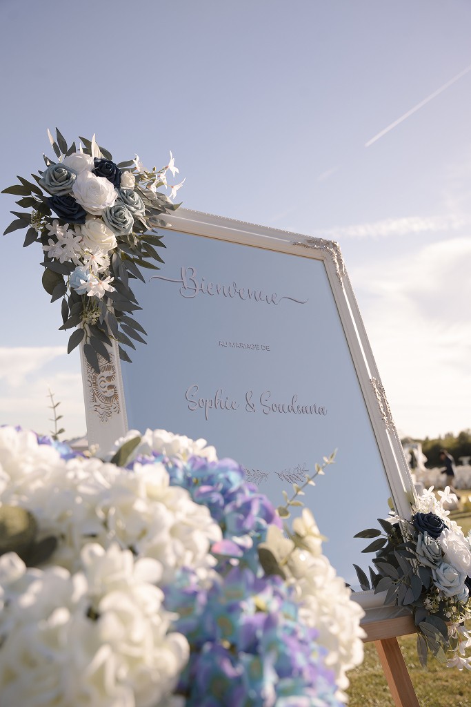 Wedding welcome sign in a white mirror frame decorated with blue and white flowers, under a clear sky