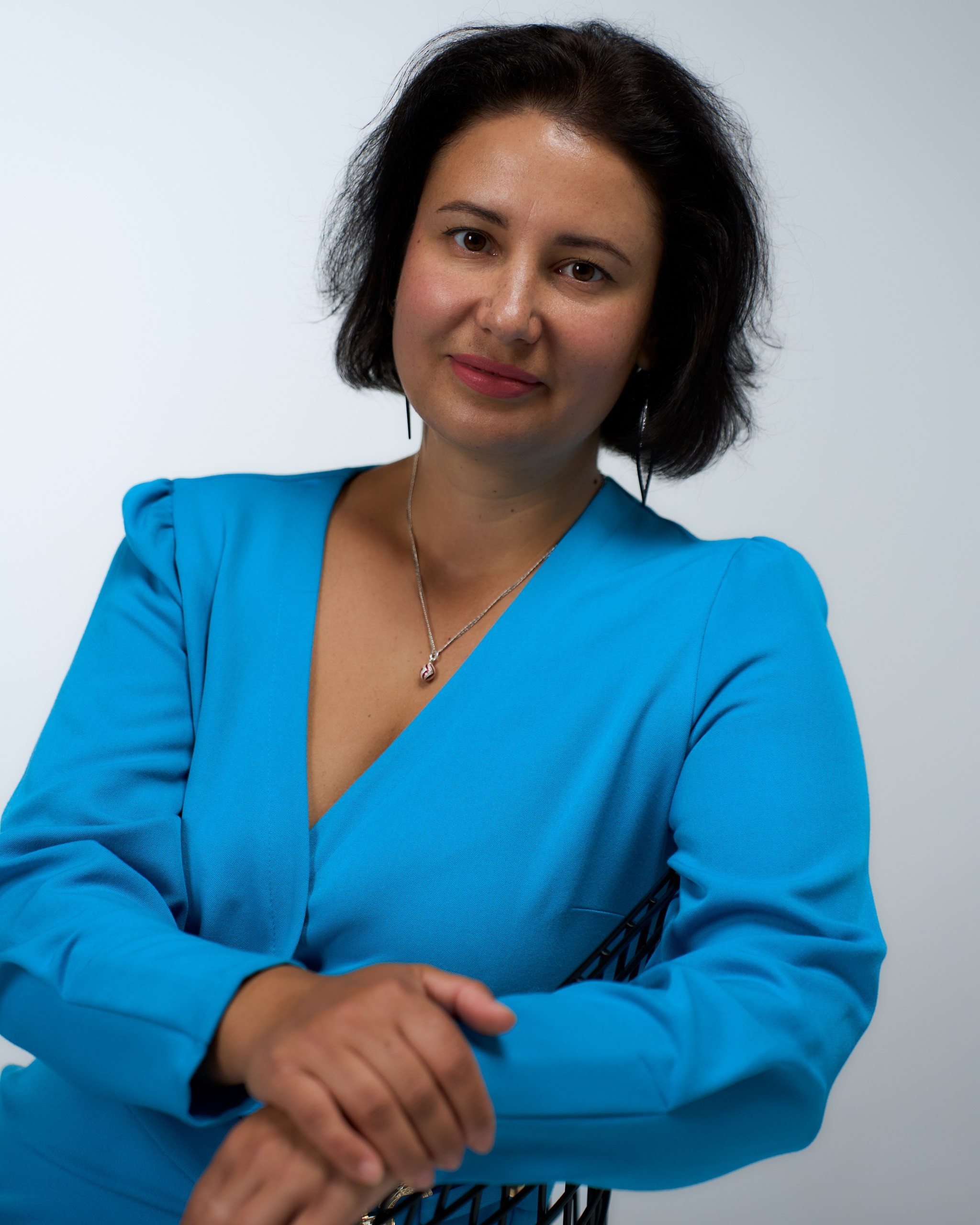 A person in a blue dress sits on a chair, looking forward, with hands clasped on the chair in front of them.