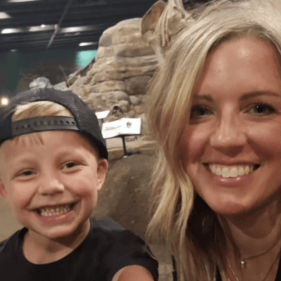 Alt tag: Portrait of Kelli Argento smiling alongside her young son wearing a backward baseball cap, both posing together indoors with a rock display in the background.