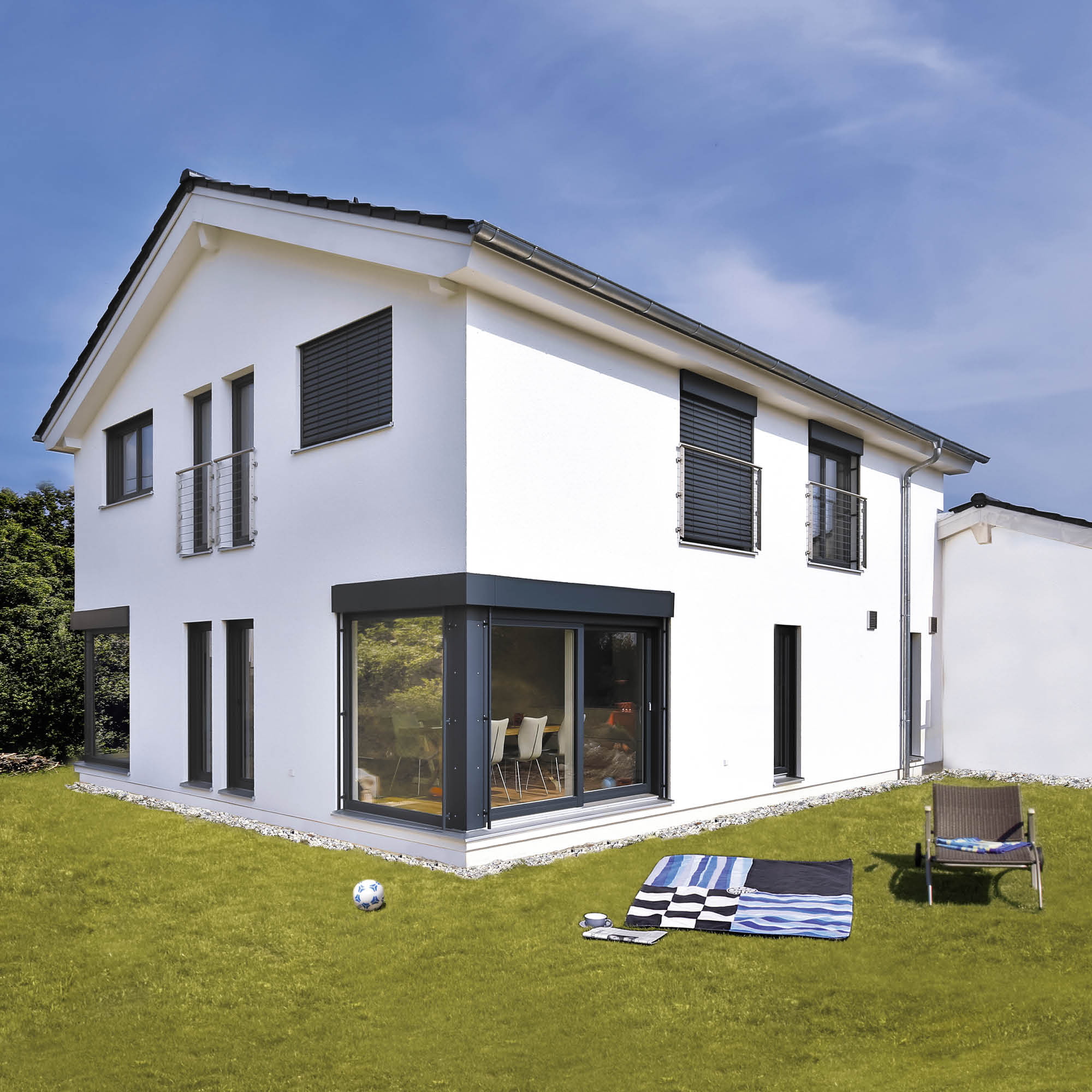 Modernes weißes Einfamilienhaus mit großzügigen Fensterfronten, Balkonen und Rollläden, Rasen, Liegestuhl und Decke im Garten.
