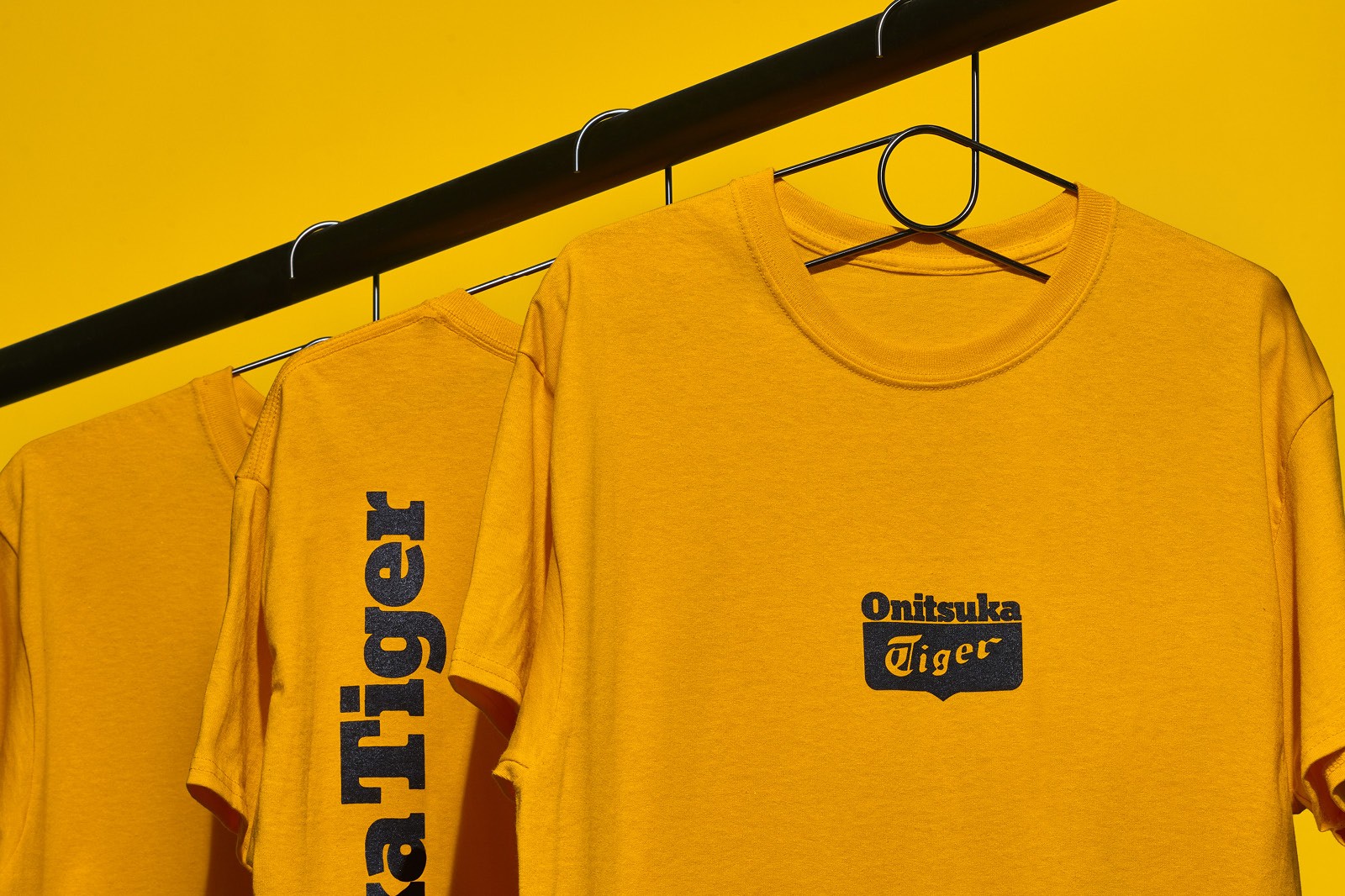 Three yellow t-shirts hang on a black hanger against a matching yellow background with logo of Onitsuka Tiger.