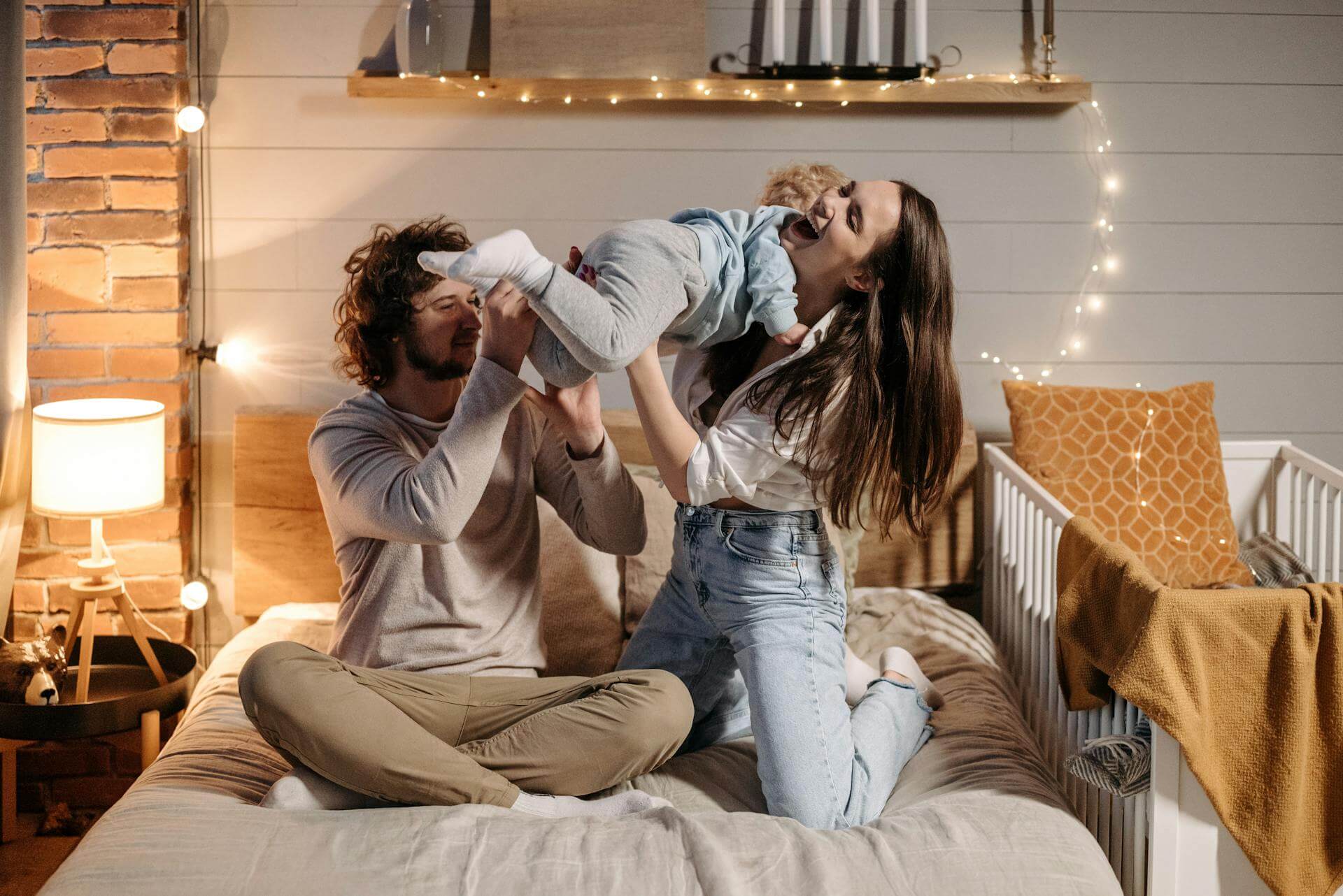 A joyful man and woman play with their baby in a cozy bedroom, surrounded by soft toys and warm lighting