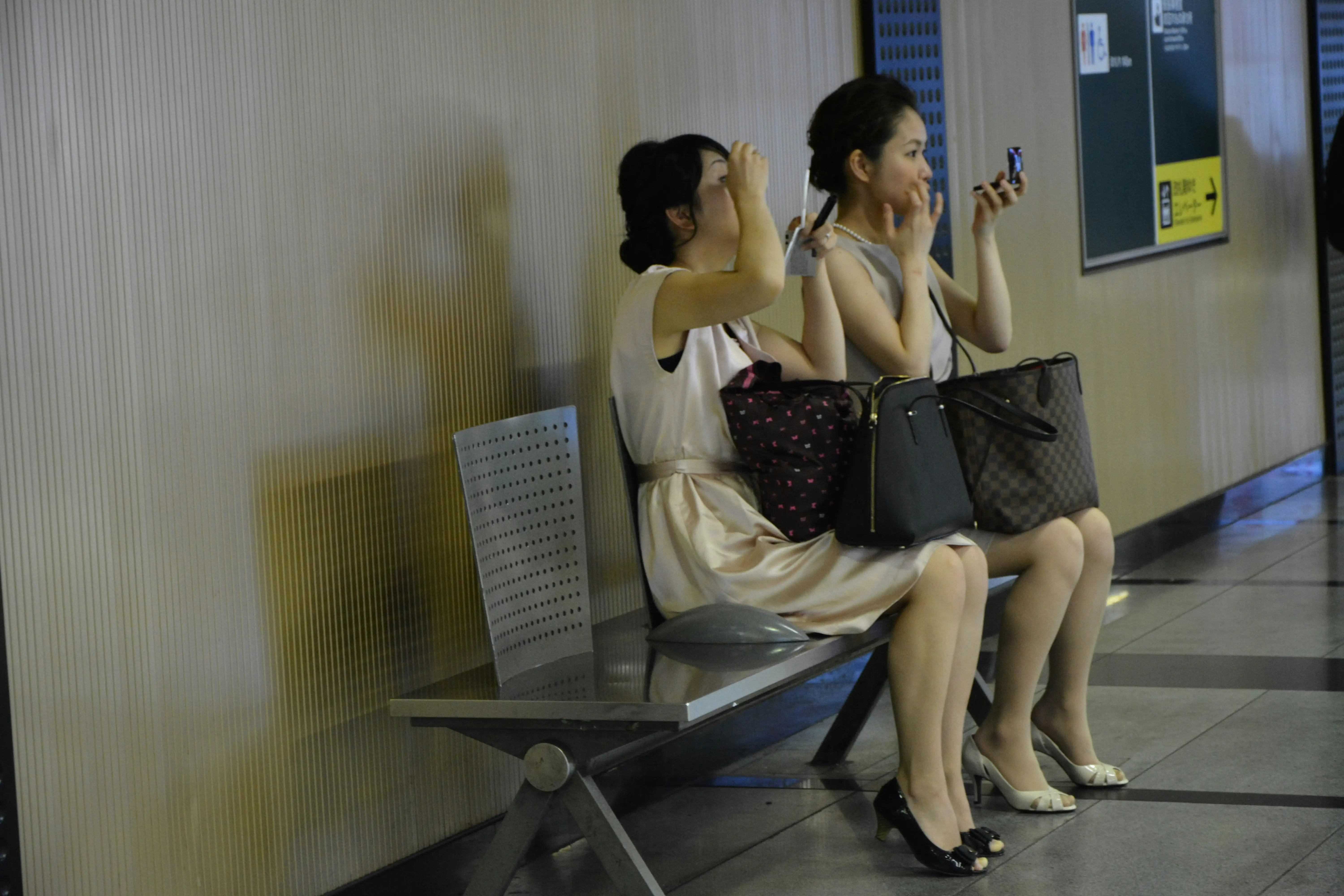 Two women sitting on a bench, chatting and using their phones, in a casual indoor setting.