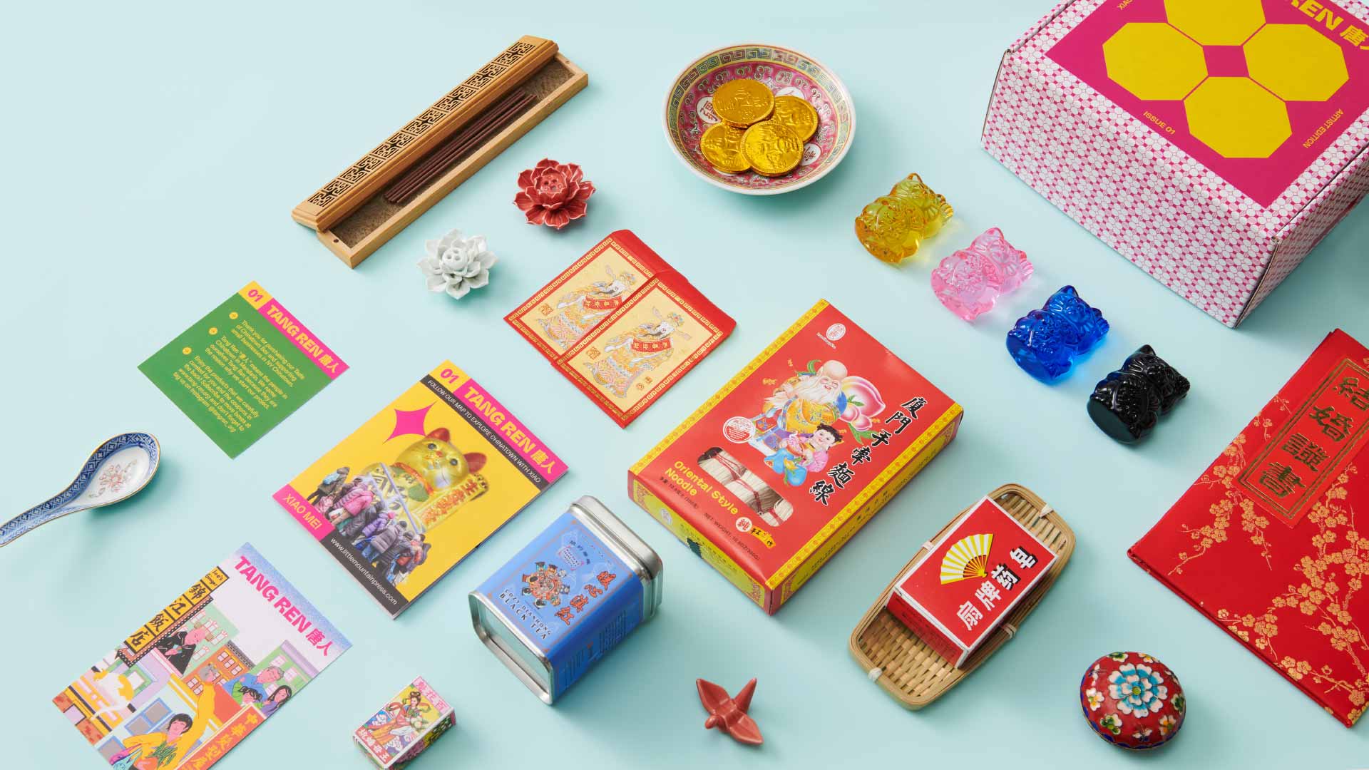 The image shows a collection of items arranged on a light blue background. These include two boxes—one red with a yellow border and Chinese characters, and another pink with a yellow geometric pattern—both featuring Chinese New Year imagery. A white bowl with gold coins, a blue bowl with black beads, a small white flower, a red rose, and a small blue container are also present. A blue spoon, a yellow card with Chinese characters, a red booklet with illustrations, a small blue tin, and a red paper clip are also visible. A red envelope with Chinese characters and a red decorative box with gold details are also included in the arrangement.