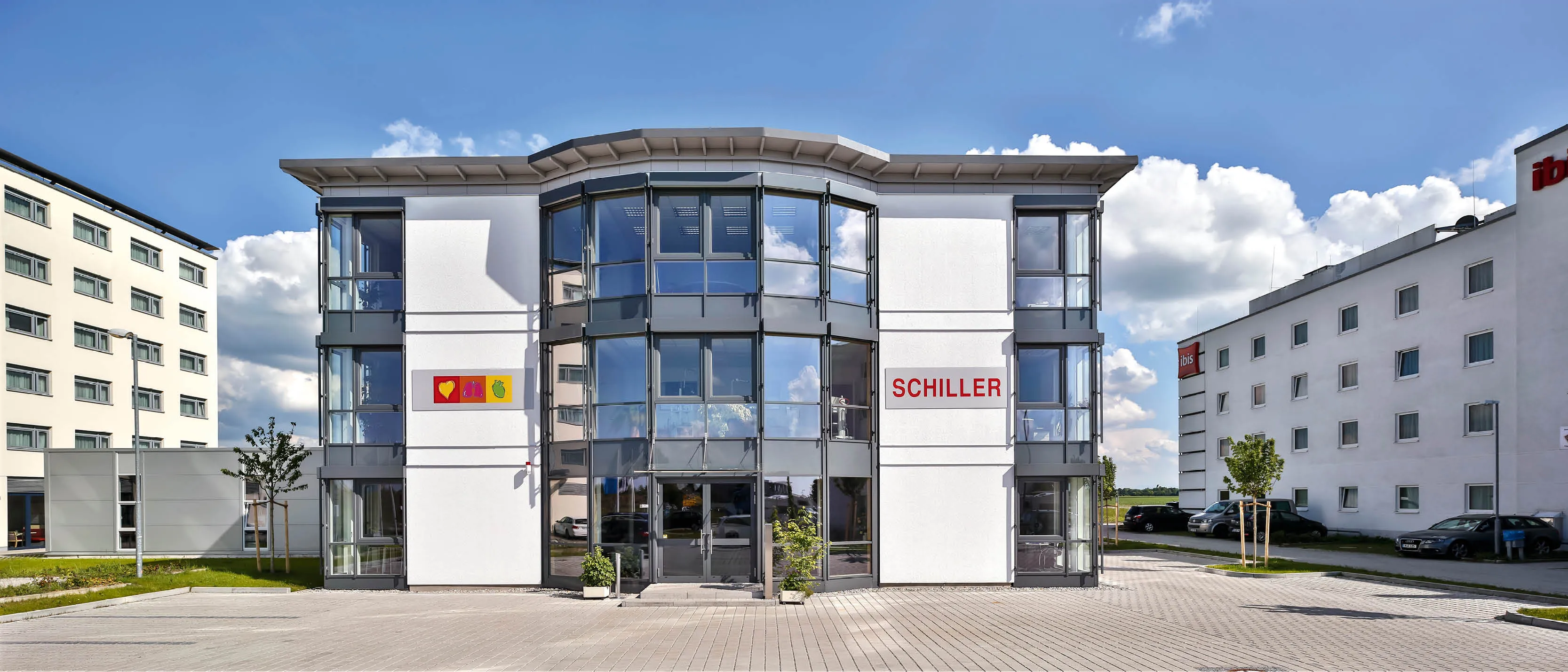 Modernes Bürogebäude mit Glaselementen und Schildern "Schiller" sowie angrenzendem Hotelgebäude.