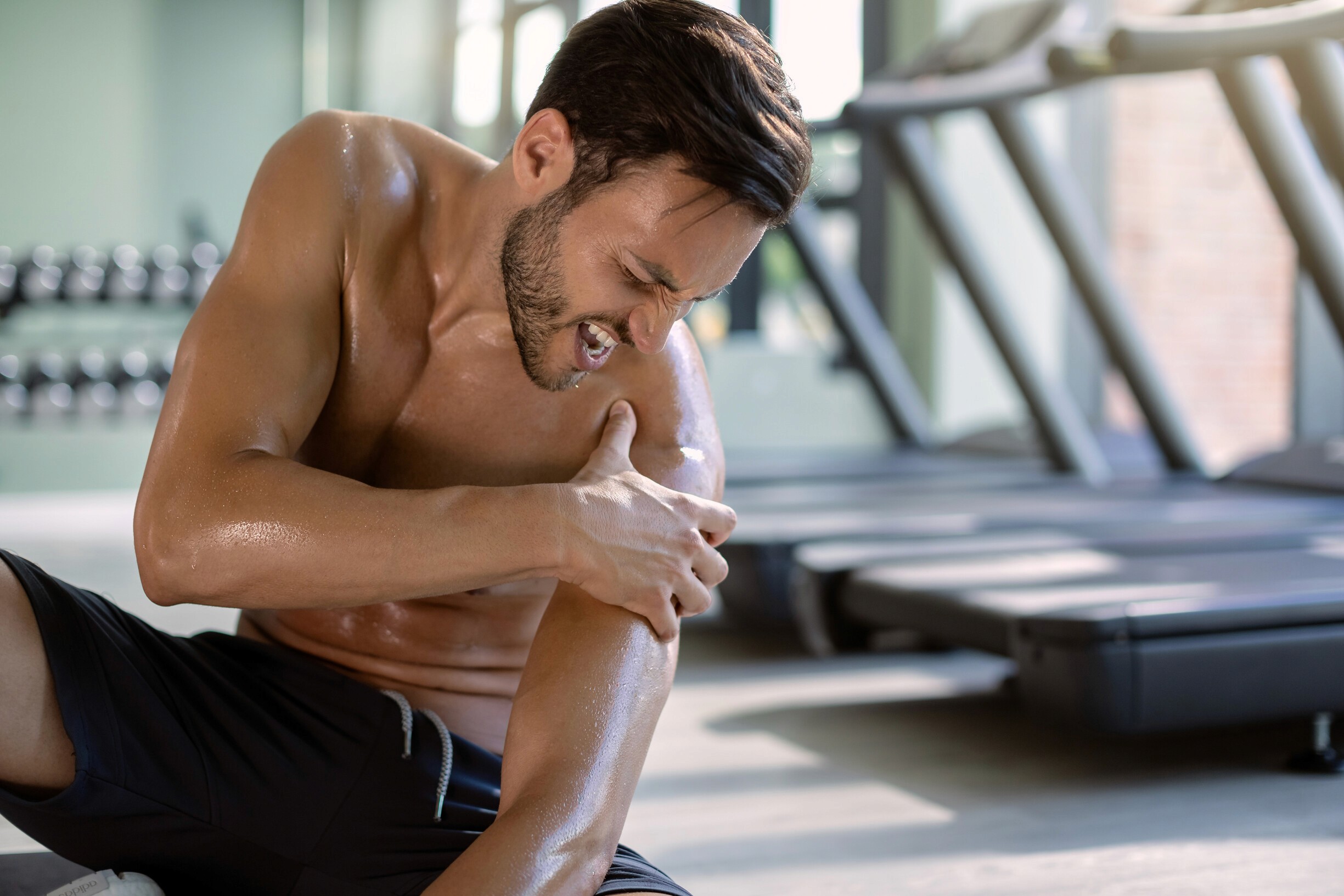 Shirtless man in a gym grimaces in pain, holding his shoulder. Background shows treadmills and weights, conveying a workout injury.