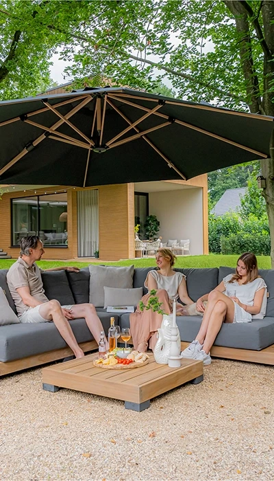 A modern outdoor seating area features three people relaxing under a large umbrella, surrounded by greenery and a contemporary house.