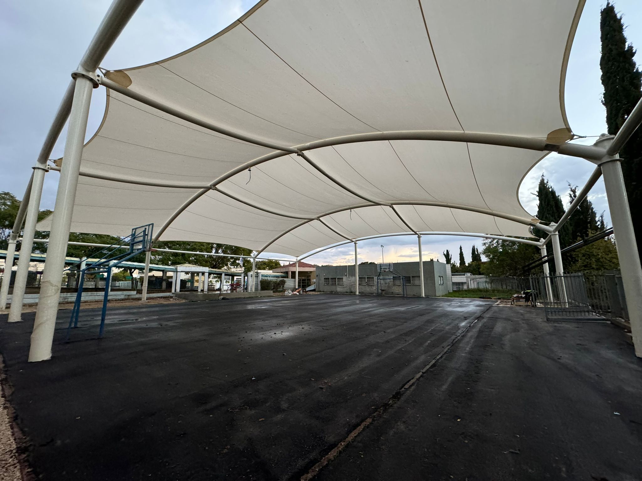 Large beige curved fabric canopy over an asphalt court with a basketball hoop and trees in the background.
