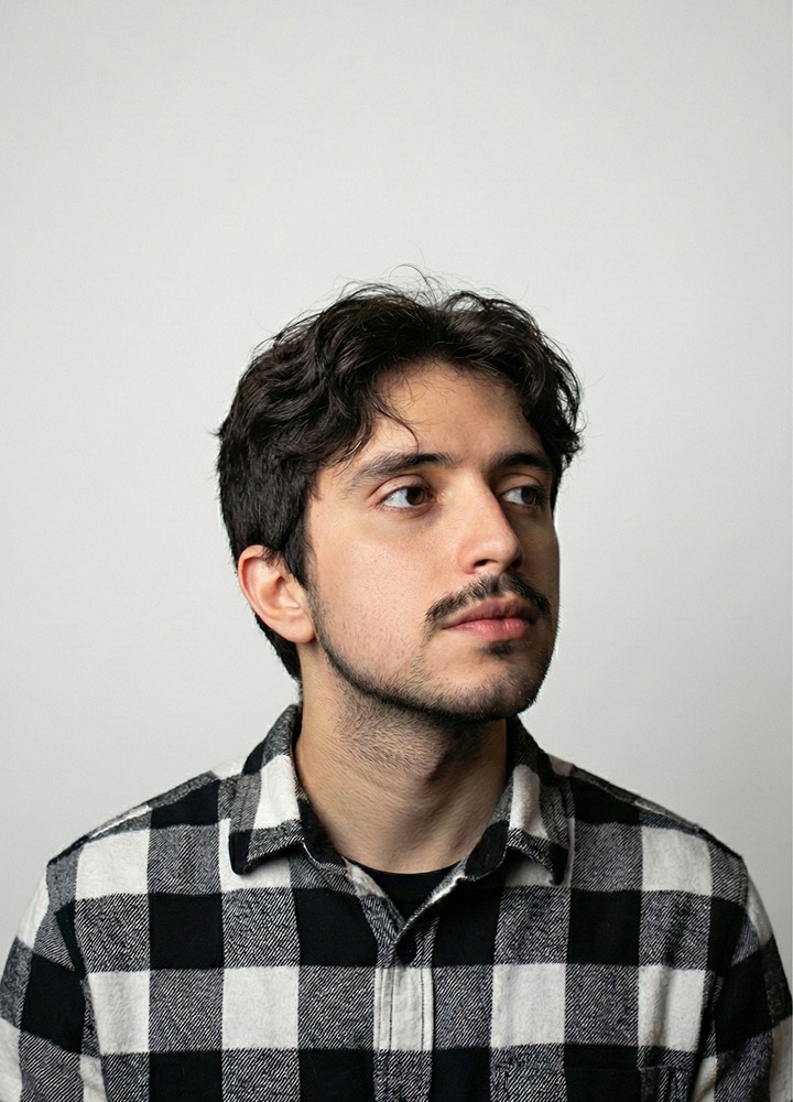 Retrato de um homem jovem de pele clara, posicionado de frente com o rosto virado levemente para a direita. Ele possui cabelos castanhos escuros, ondulados e curtos, além de bigode e barba rala. Veste uma camisa de botão xadrez em preto e branco sobre uma camiseta preta básica, sem o uso de acessórios visíveis. A iluminação é branca e suave, característica de estúdio profissional, incidindo sobre o rosto contra um fundo azul sólido.