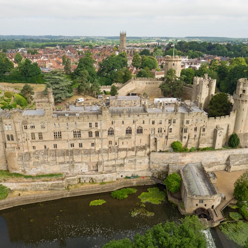 Ilmakuva suuresta kivilinnasta, jossa on useita torneja, ympärillä vehreää luontoa, vettä ja kaukainen kaupunkimaisema.