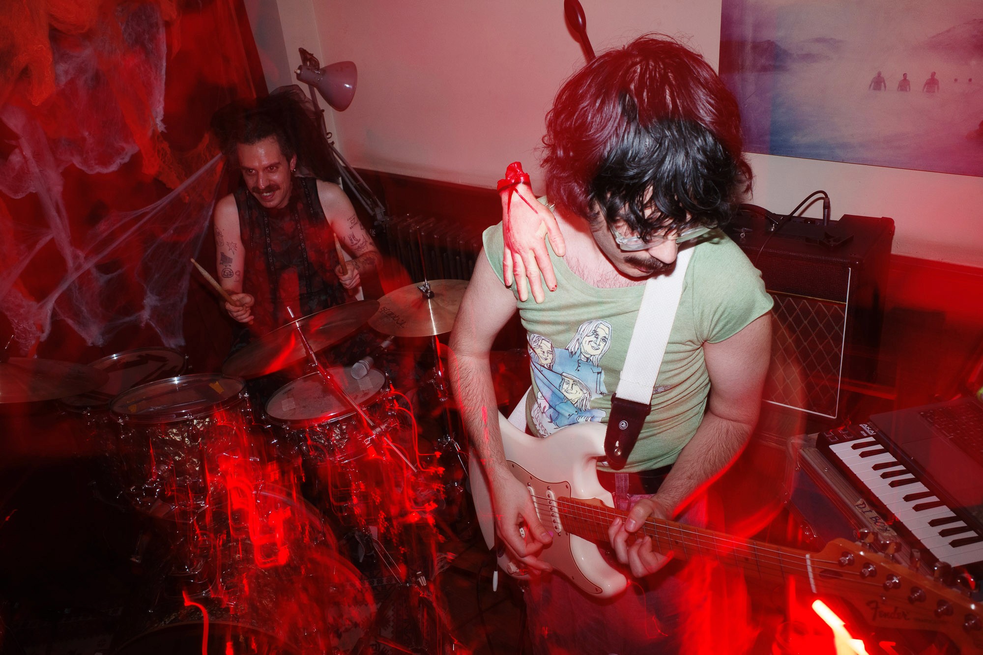 A guitarist passionately plays an electric guitar with a red light effect, while a drummer enthusiastically performs on a drum kit in a dimly lit room, creating a vibrant live music atmosphere.