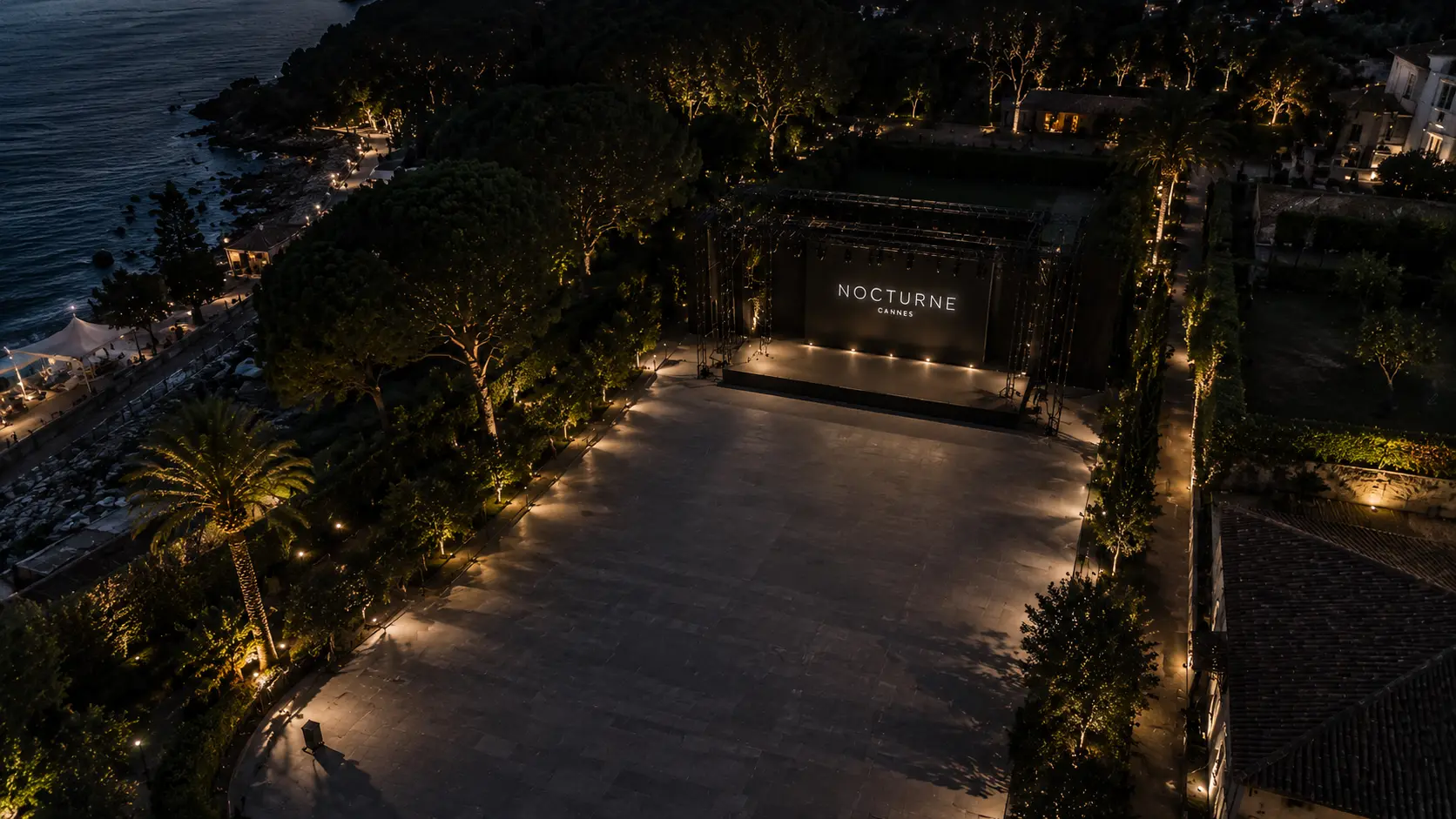 Vue drone d’une scène de défilé outdoor haut de gamme sur la Côte d’Azur au crépuscule, avec scénographie élégante et décors illuminés.