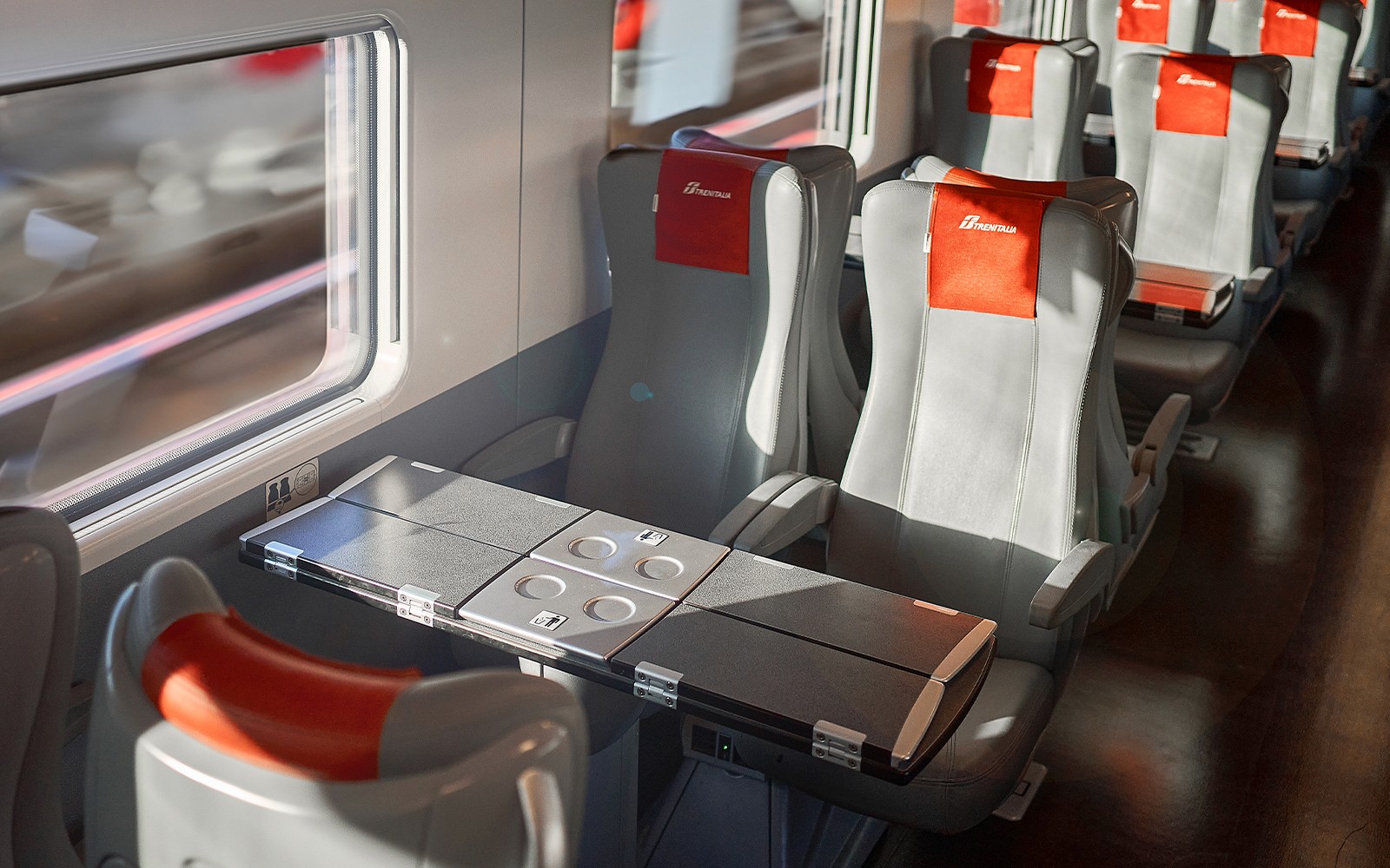 Trenitalia train standard class seats with foldable tables and window view.