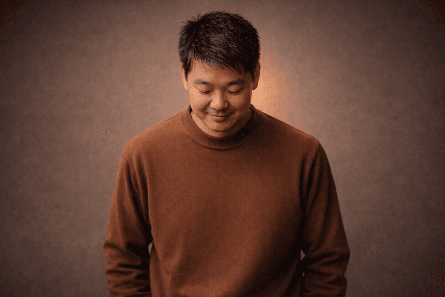 A person in a brown sweater stands against a warm, softly lit gray background, looking down with a slight smile.
