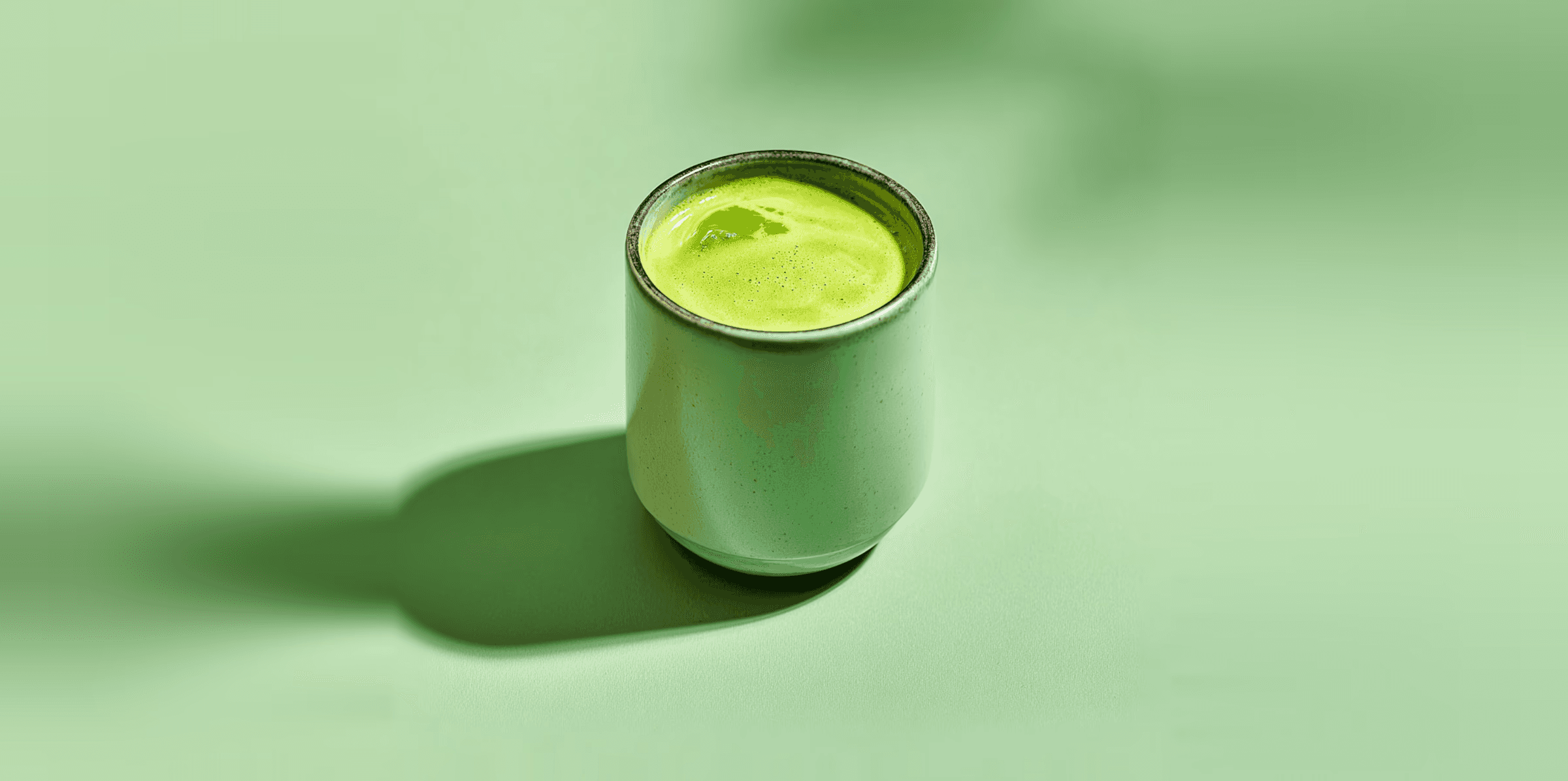 Minimalist still life of a matcha green drink in a ceramic cup on a pale green surface with long artistic shadows.
