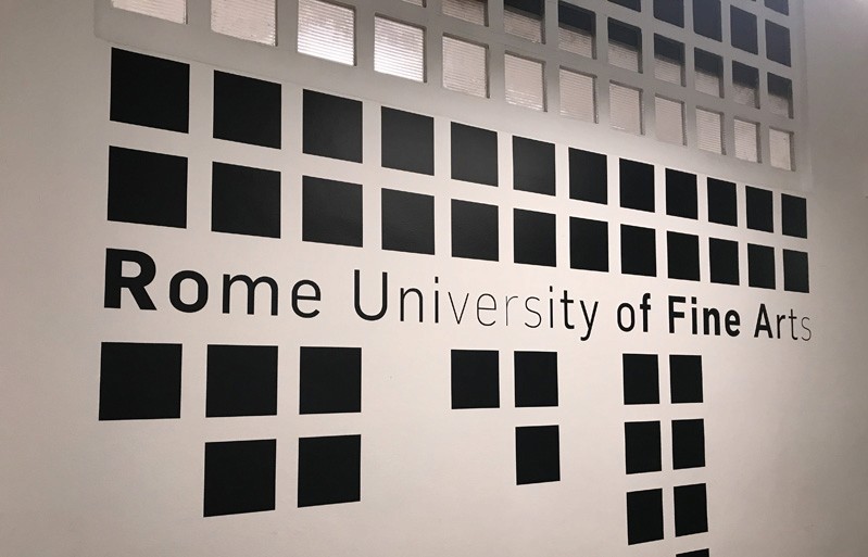 A picture of some black squares on a white wall, as wel as the text: Rome University of Fine Arts.