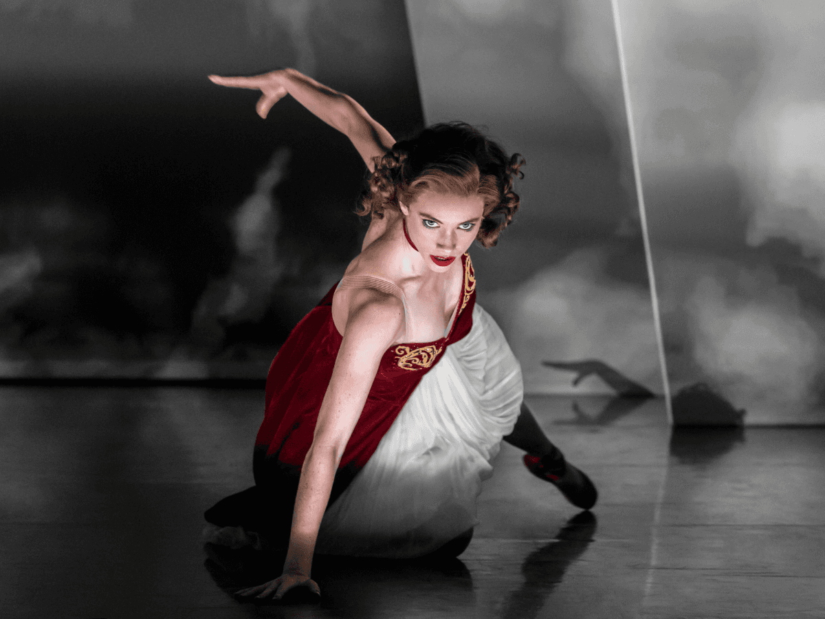 赤とクリーム色のドレスを着たソロダンサーが、スポットライトの中で動きの途中でしゃがみ込んでいます。