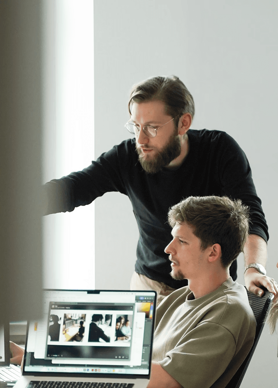 Two men collaborate in a bright office; a bearded man in glasses points at a laptop showing photos as a colleague watches.