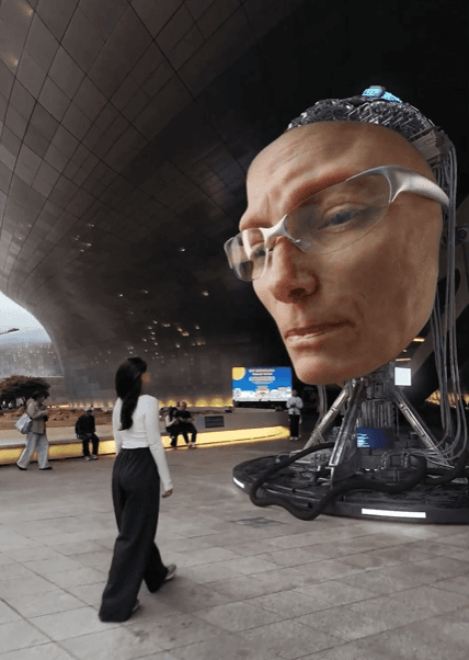 A woman walking toward a massive, futuristic Tilda Swinton robotic head in a modern city plaza