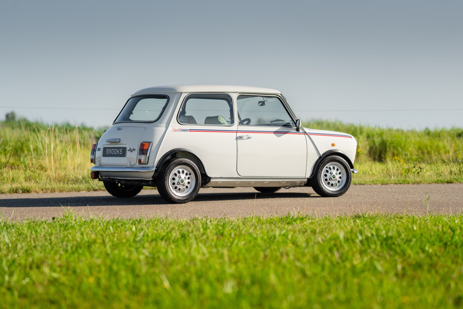 A classic white car parked on a road surrounded by green grass and trees under a clear blue sky.