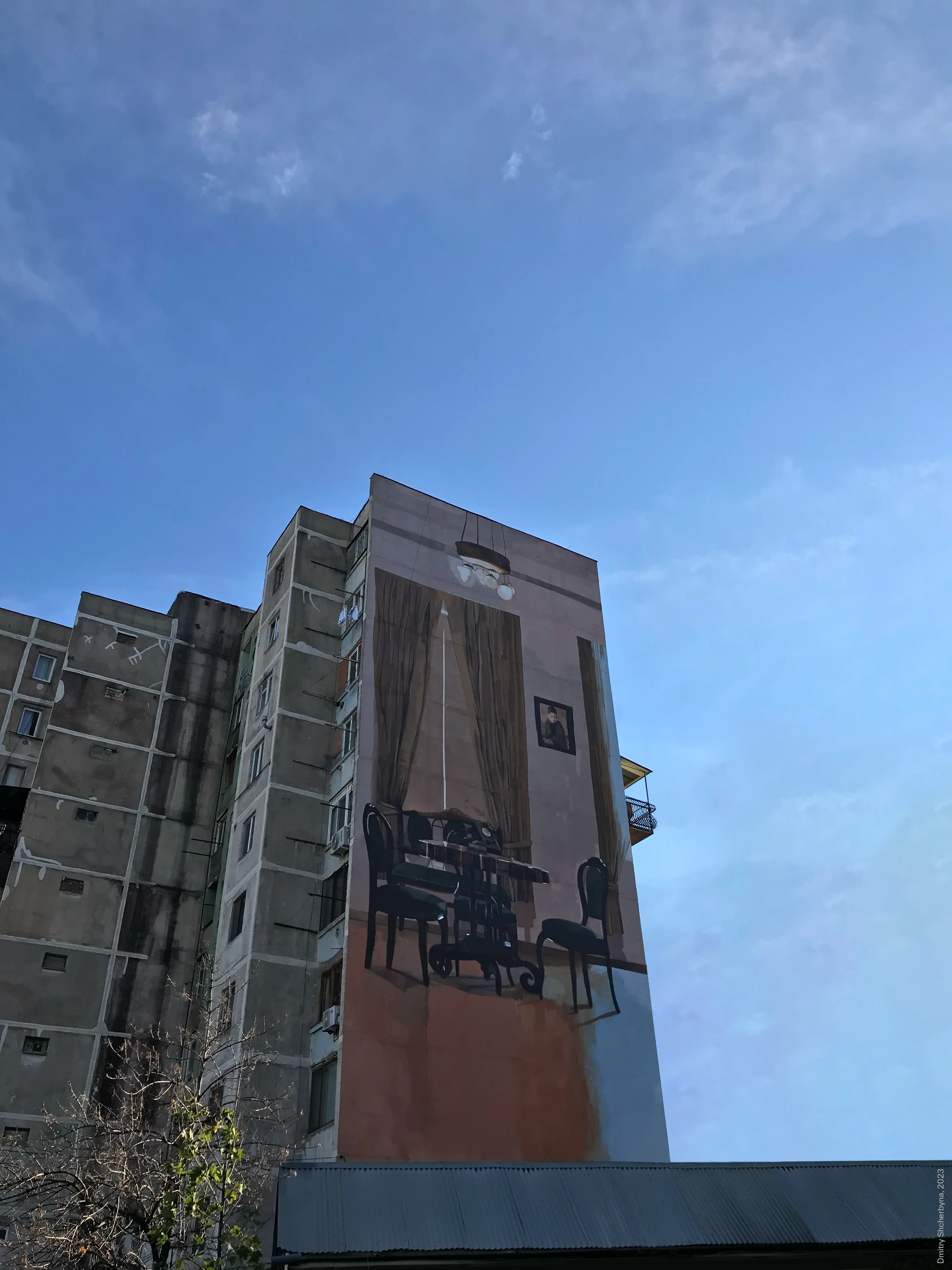 A mural on the side of a high-rise apartment building depicts a traditional interior room with dark wooden furniture, a table with chairs, long curtains flanking a bright window, a framed portrait on the wall, and a hanging light fixture. The painting creates the illusion of a room on the building's exterior.