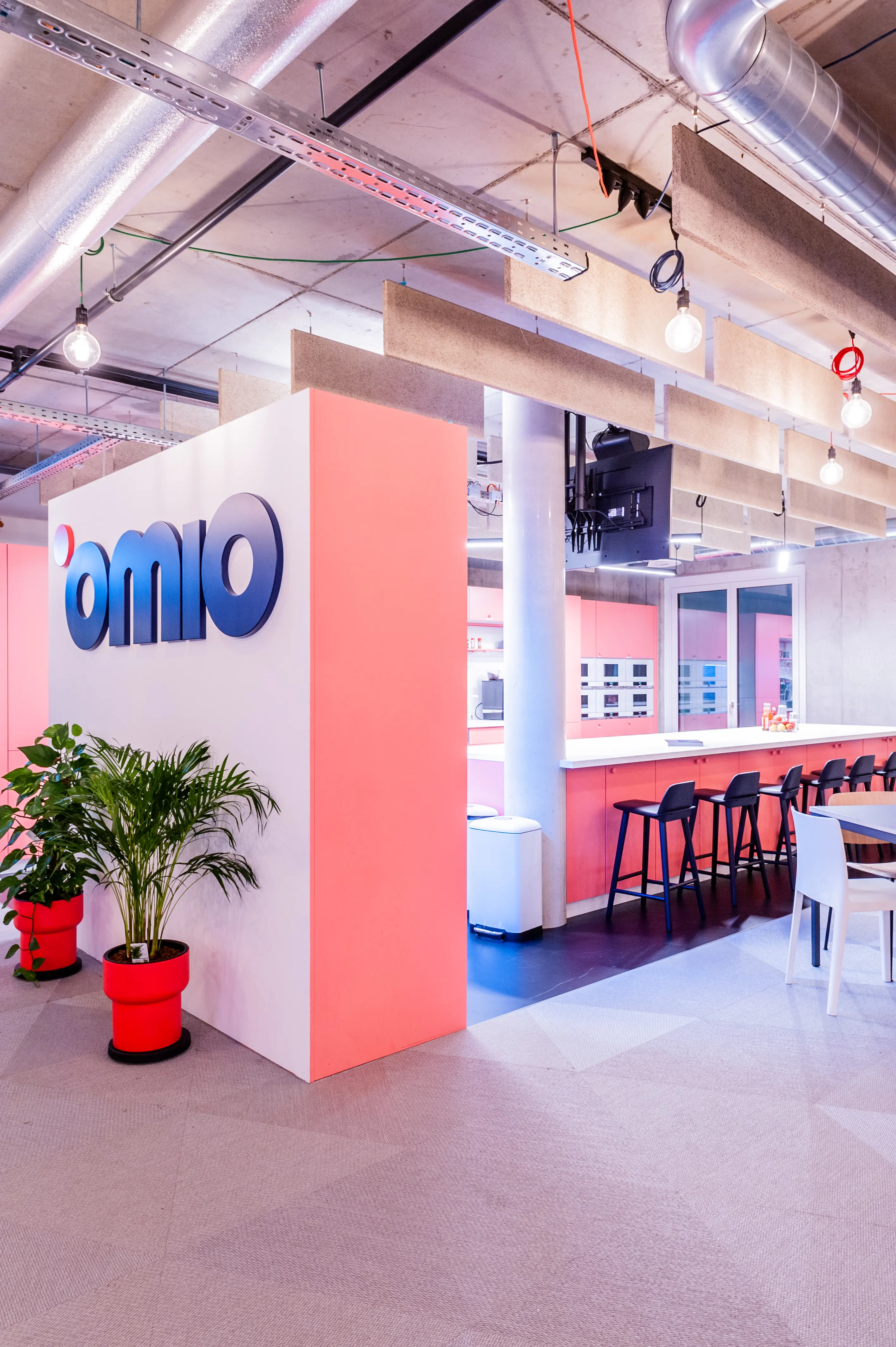 Coral-pink kitchen bar at OMIO with black bar stools, a white wall block with the blue Omio logo, palm plants in red pots and exposed ventilation ducts above.