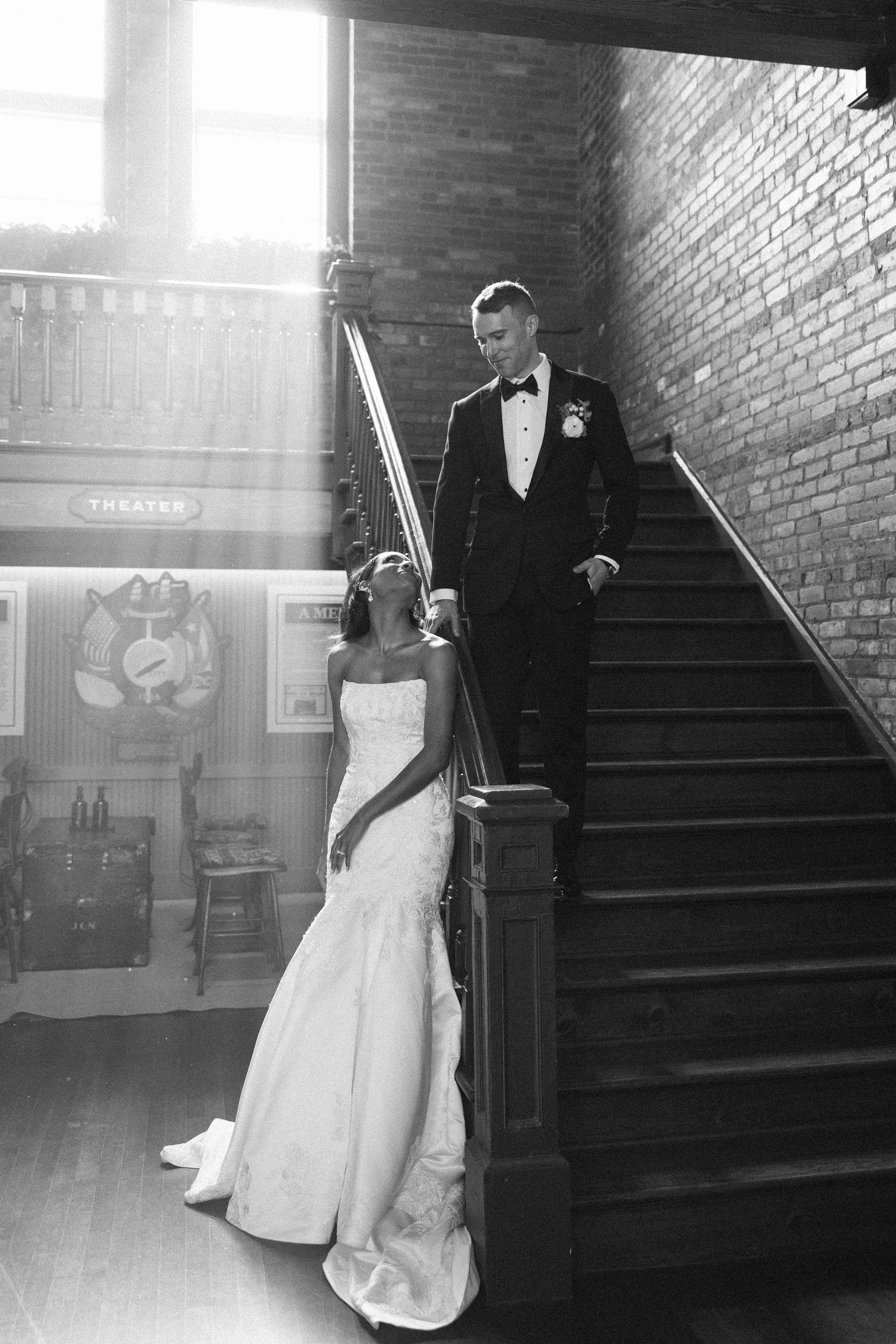 A black-and-white photo of a wedding couple walking down a staircase. The bride is wearing a white dress and the groom is in a tuxedo.