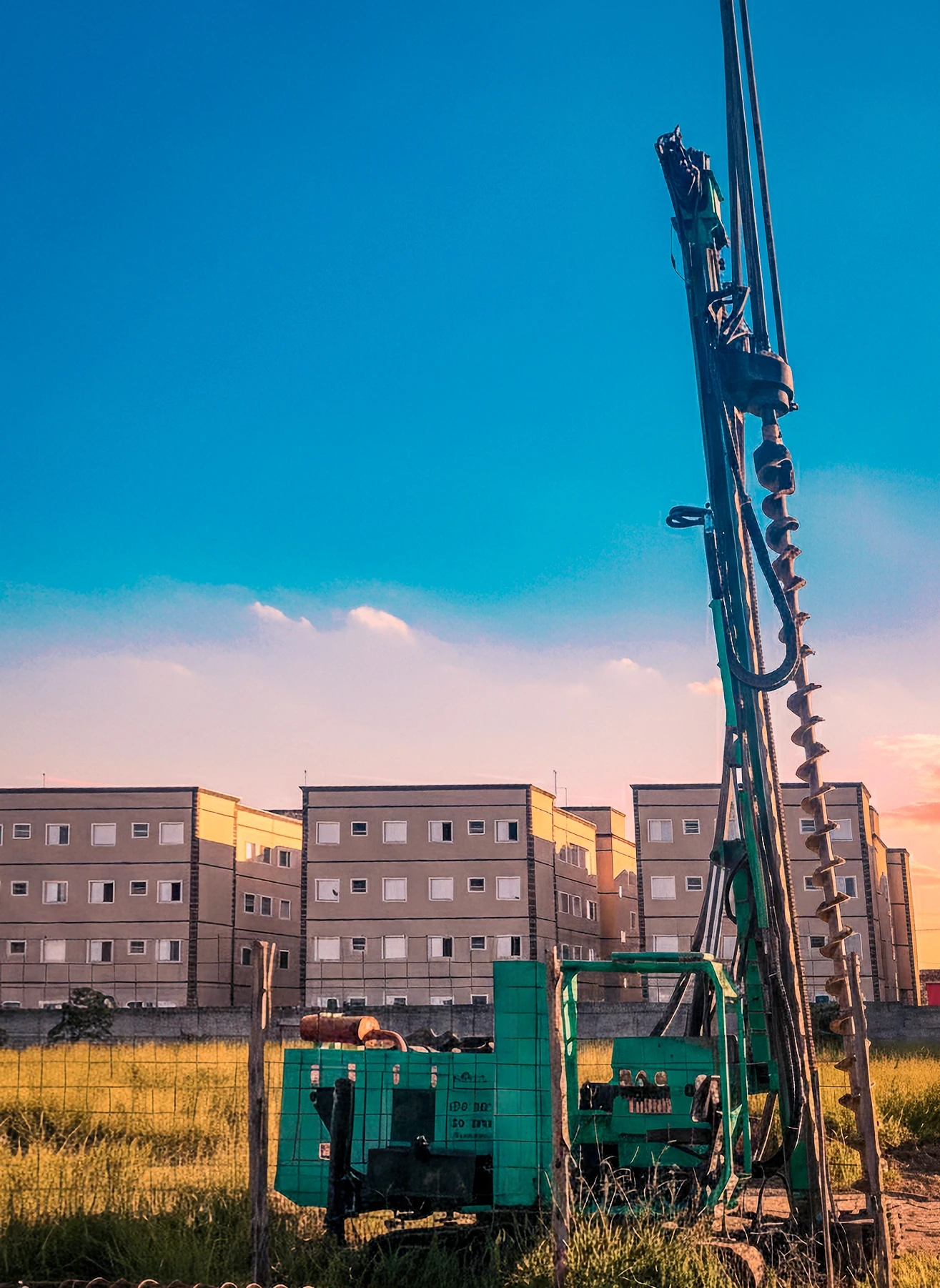 Máquina de perfuração em primeiro plano, com o trado de hélice em destaque, em um campo de grama. Ao fundo, vários edifícios residenciais grandes e idênticos (apartamentos) sob um céu azul.