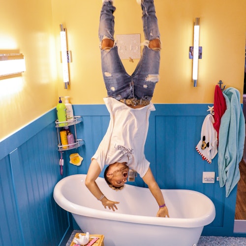 Person doing a handstand in a bathroom, with blue paneling and a white bathtub. Accessories and towels are on the walls.