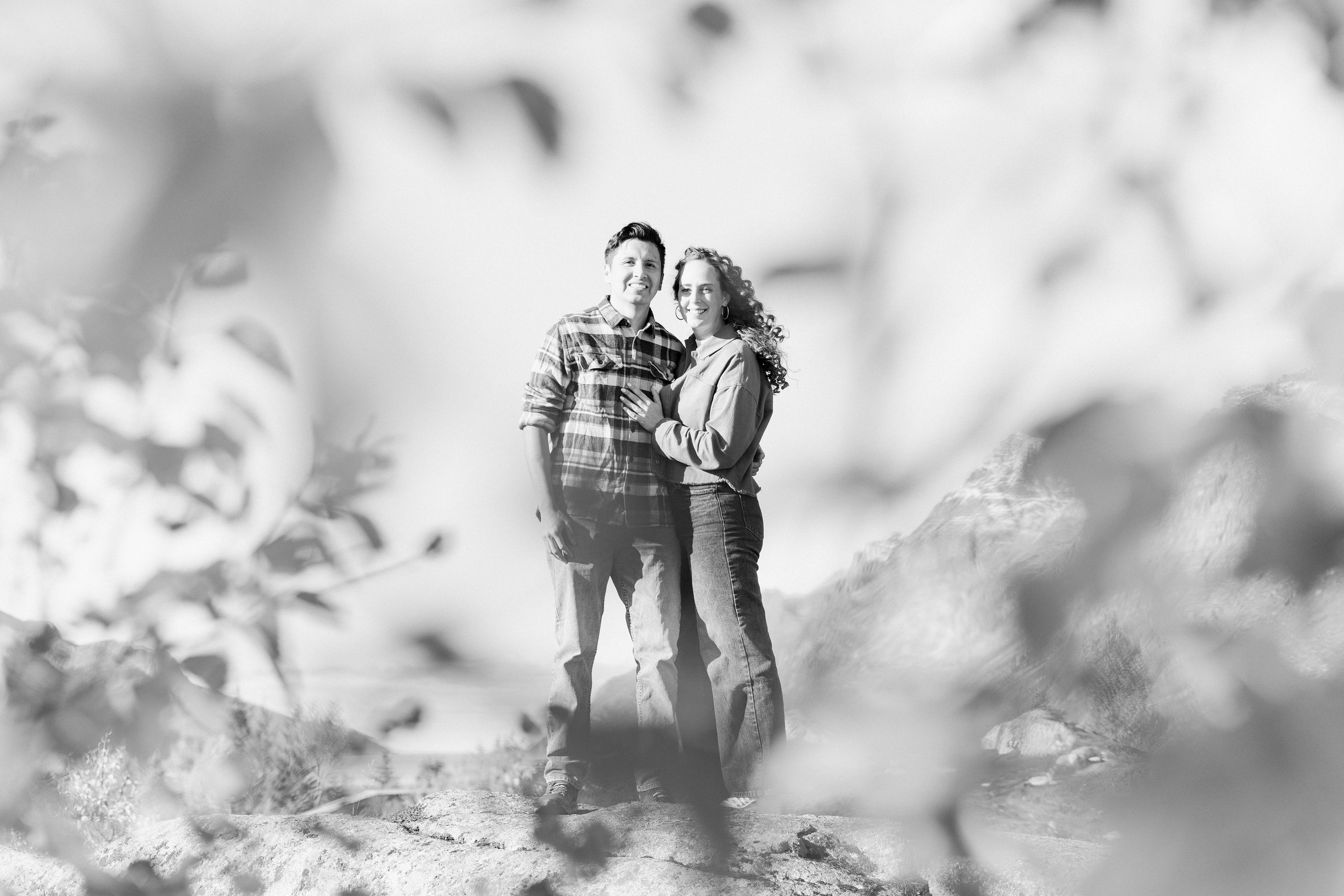 In black and white, the couple stands together behind softly blurred foreground foliage.