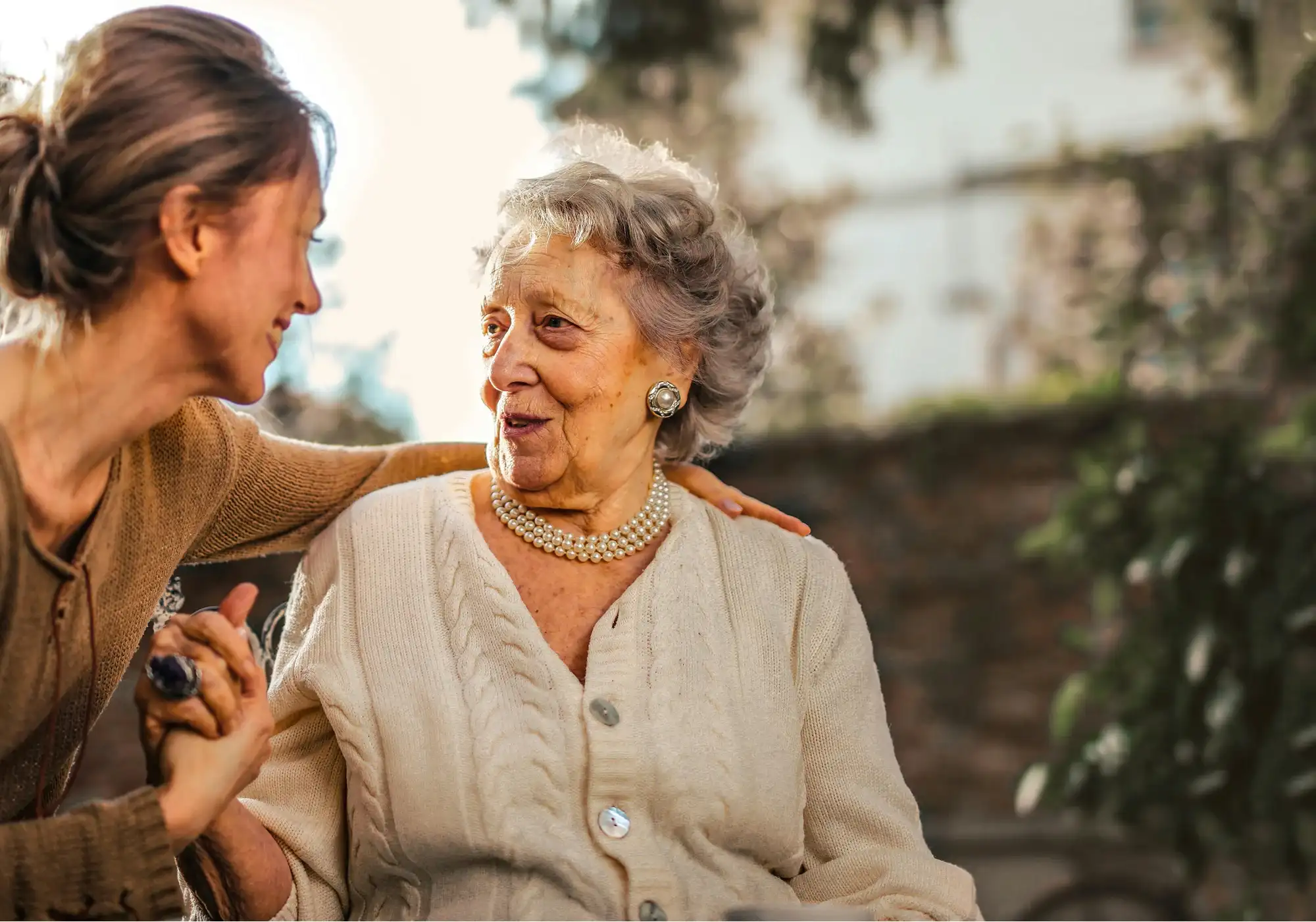 A young woman and an elderly woman sit outside, smiling warmly at each other. The young woman gently holds the other's hand. The atmosphere is tender and caring.