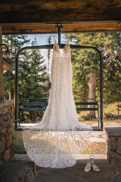 The lace wedding gown hangs from a ski-lift chair beneath the cabin porch, with bridal shoes below.