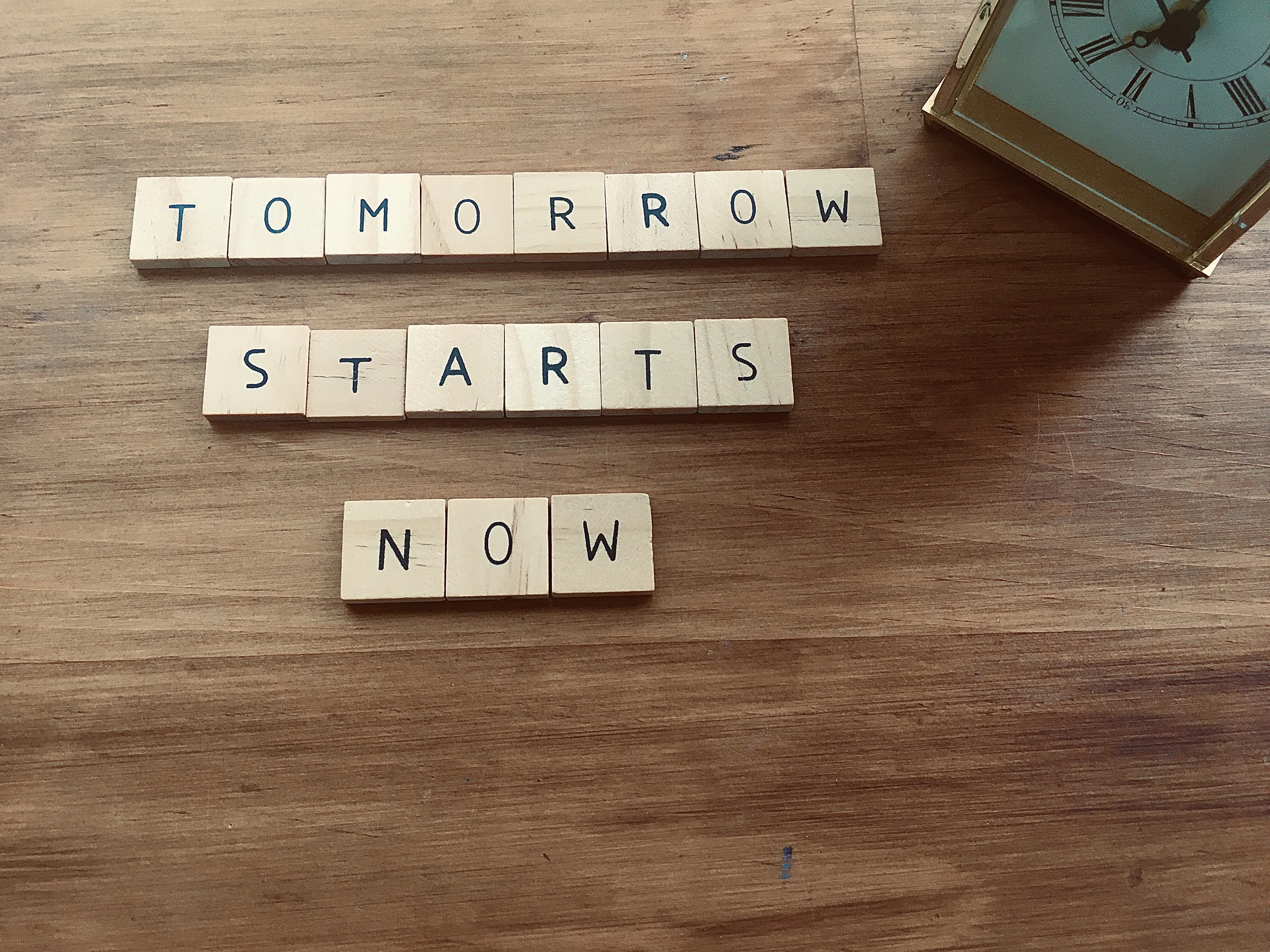 Wooden letter tiles spelling Tomorrow Starts Now next to a clock on a wooden surface, representing the urgency of getting the right conditions in place so a marketing system can start compounding at the right time
