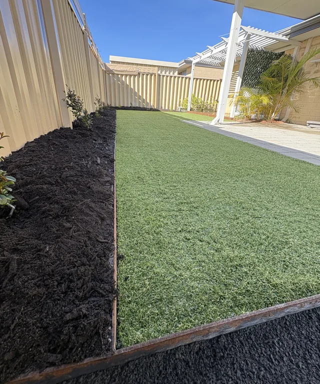 Synthetic lawn bordered by dark mulch beds and steel edging beside a paved patio.