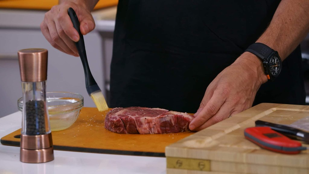 Chef seasoning ribeye steak with marinade brush on cutting board.