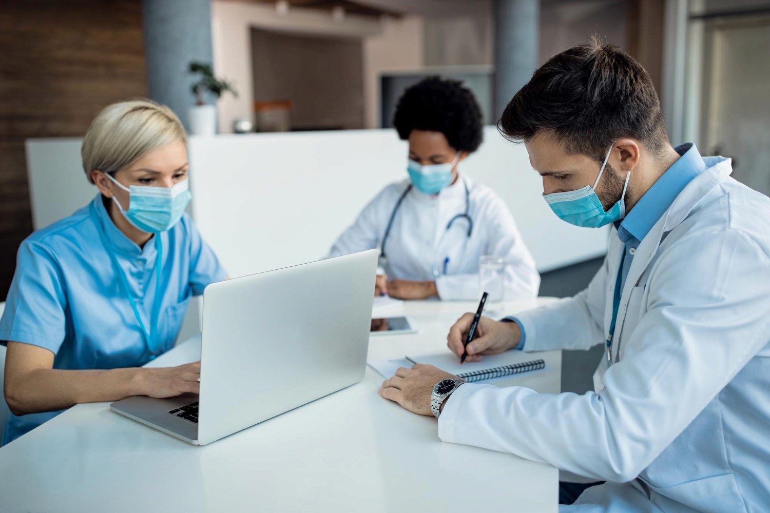 Three healthcare professionals in medical attire and face masks collaborate at a table. One uses a laptop, while the others take notes, reflecting teamwork.