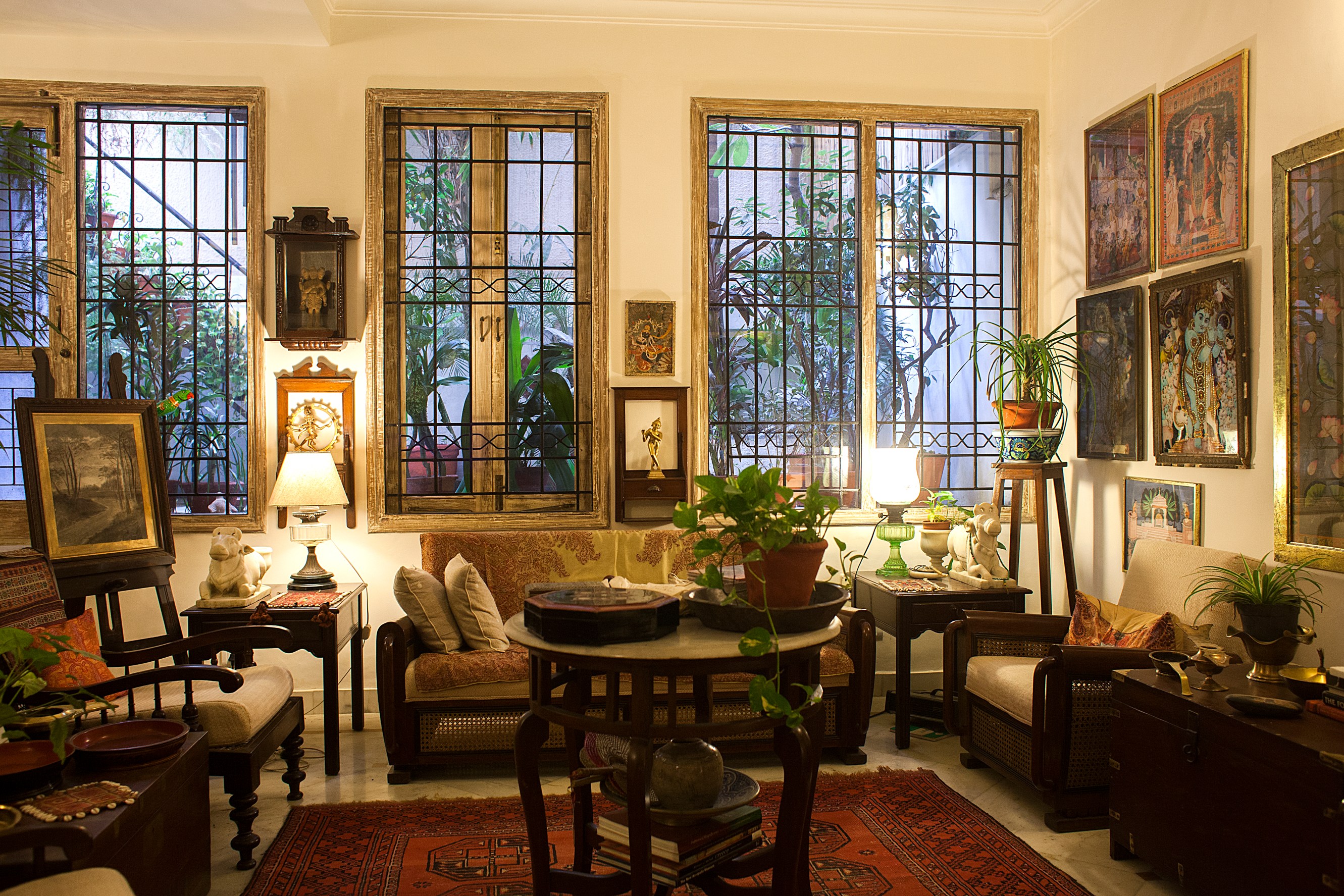 Cozy living room with warm lighting, featuring vintage furniture, framed artwork on walls, and potted plants near large windows with decorative iron grills.